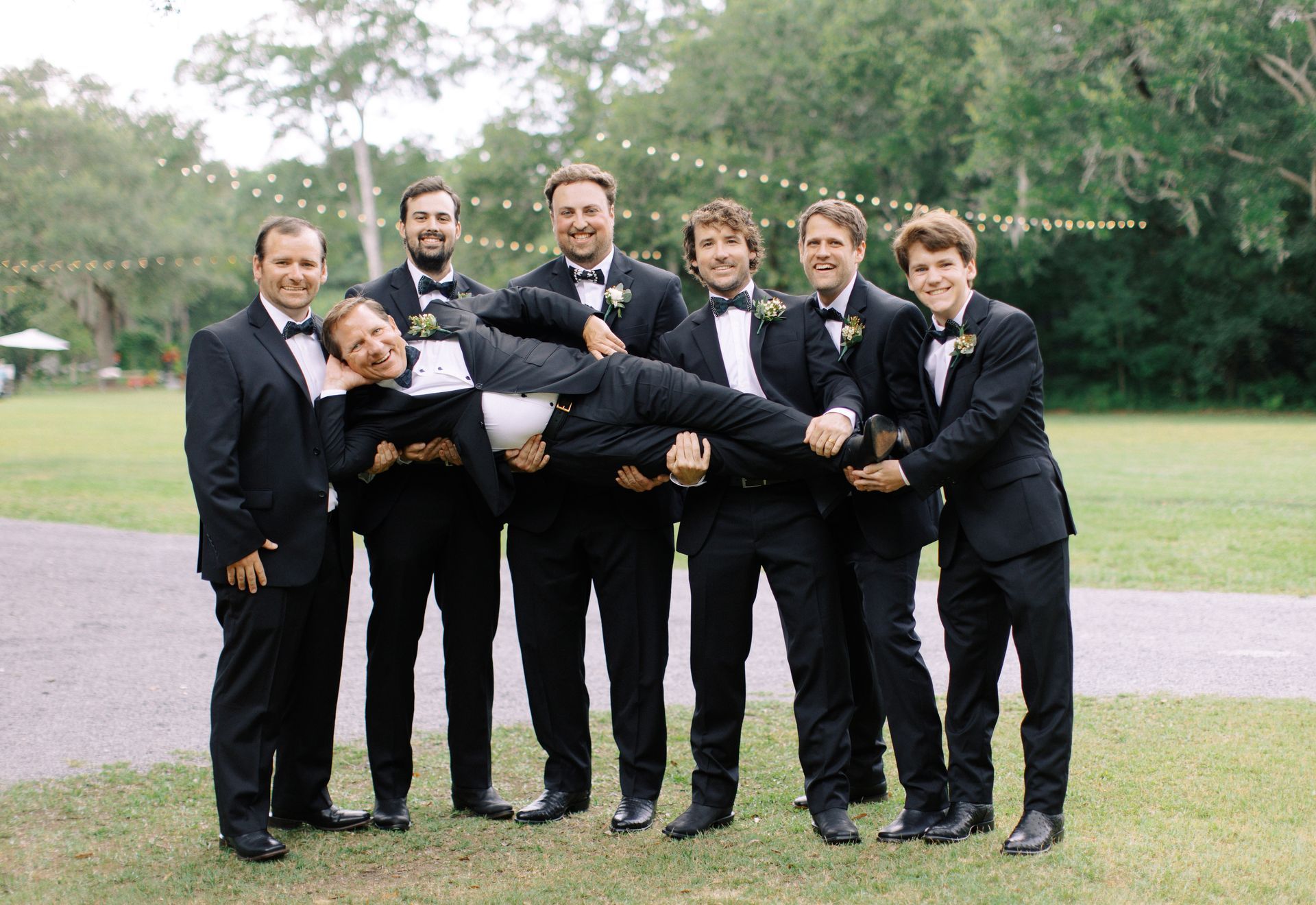 Six men in black suits posing outdoors, holding one man horizontally on a grassy lawn.