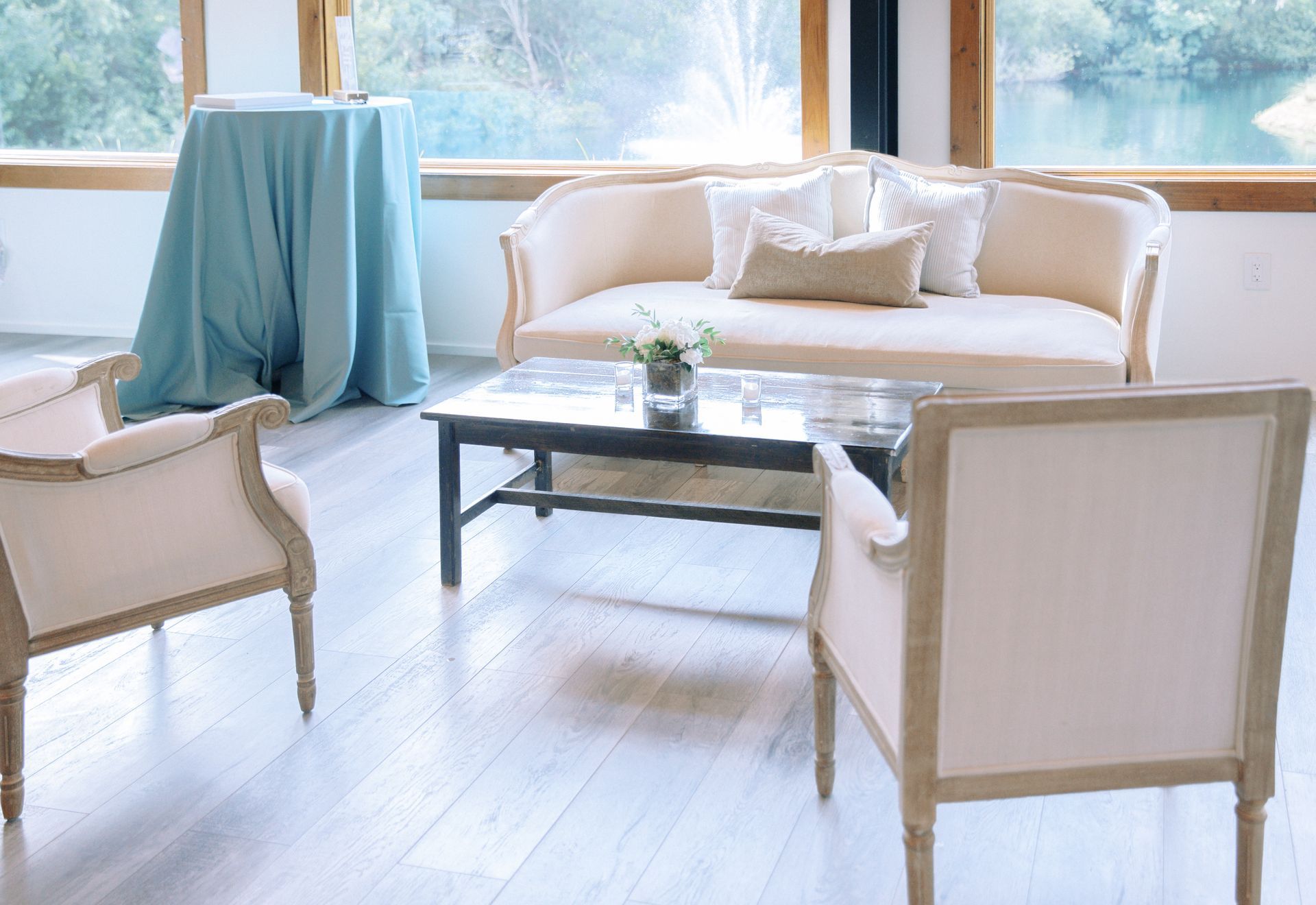 Bright living room with cream chairs, a sofa, and a glass coffee table by a sunlit window and blue water view