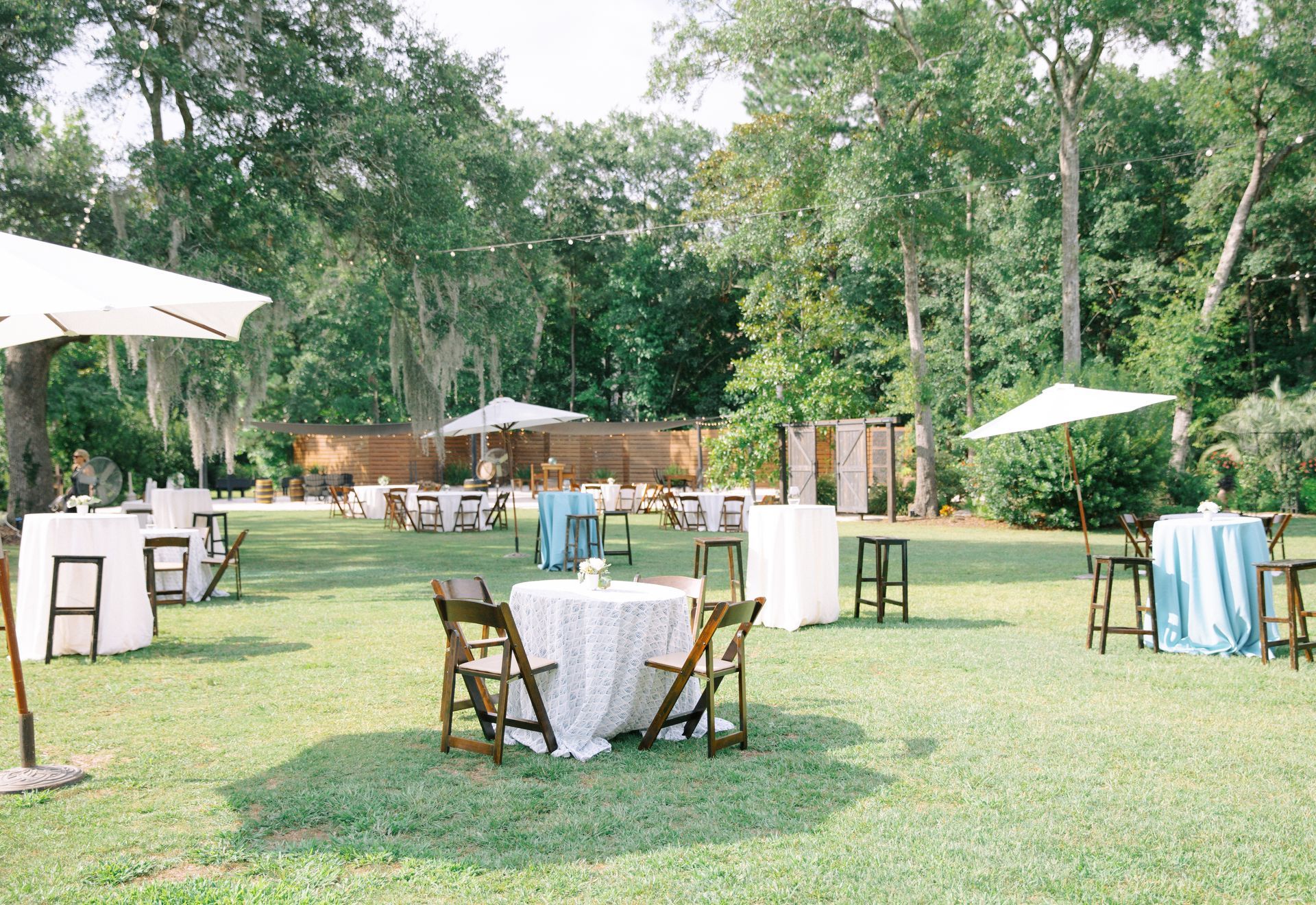 Outdoor garden event setup with white tables, chairs, umbrellas, and trees in the background