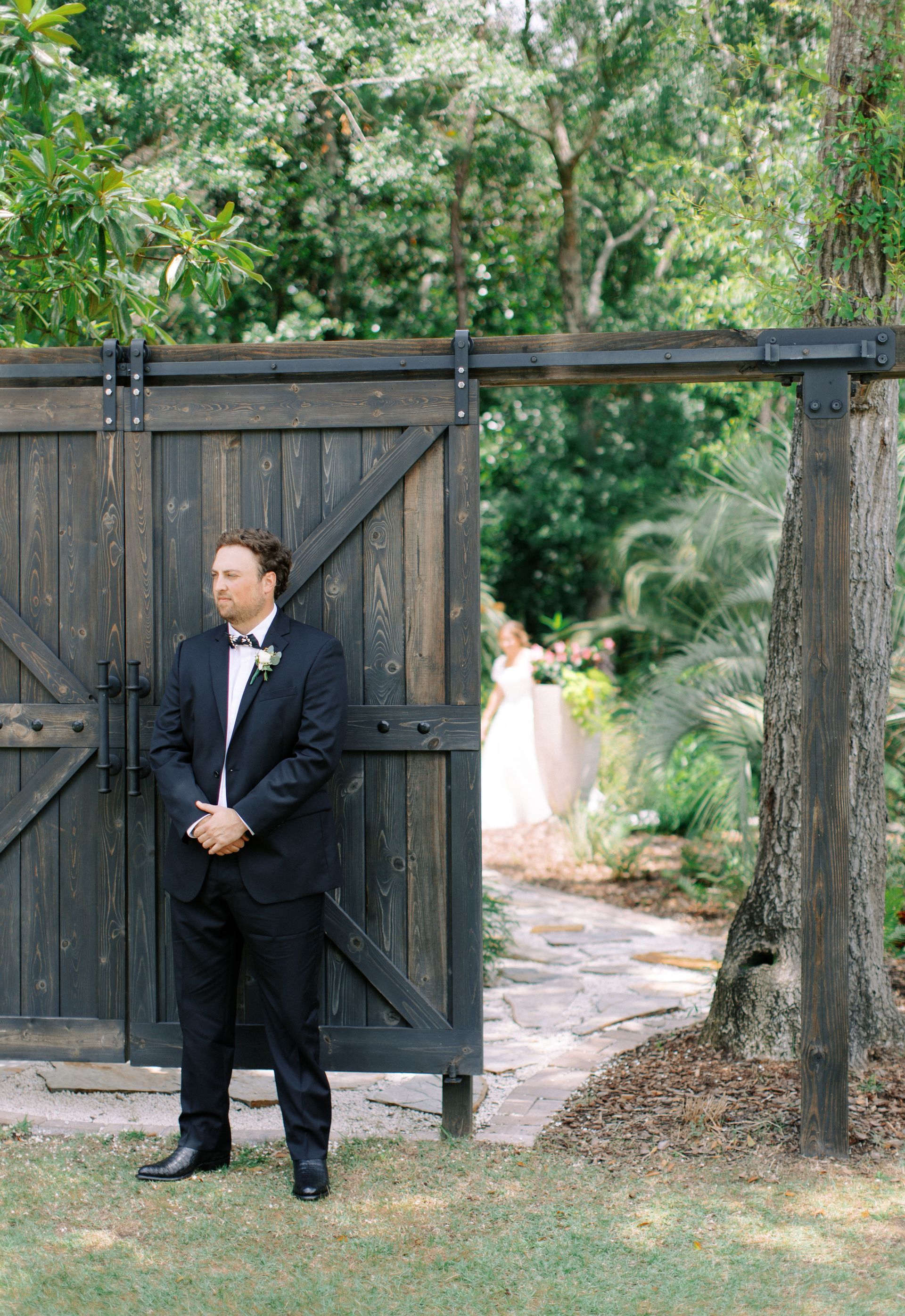Man in a dark suit stands by a wooden gate, with a bride visible on a garden path behind him.