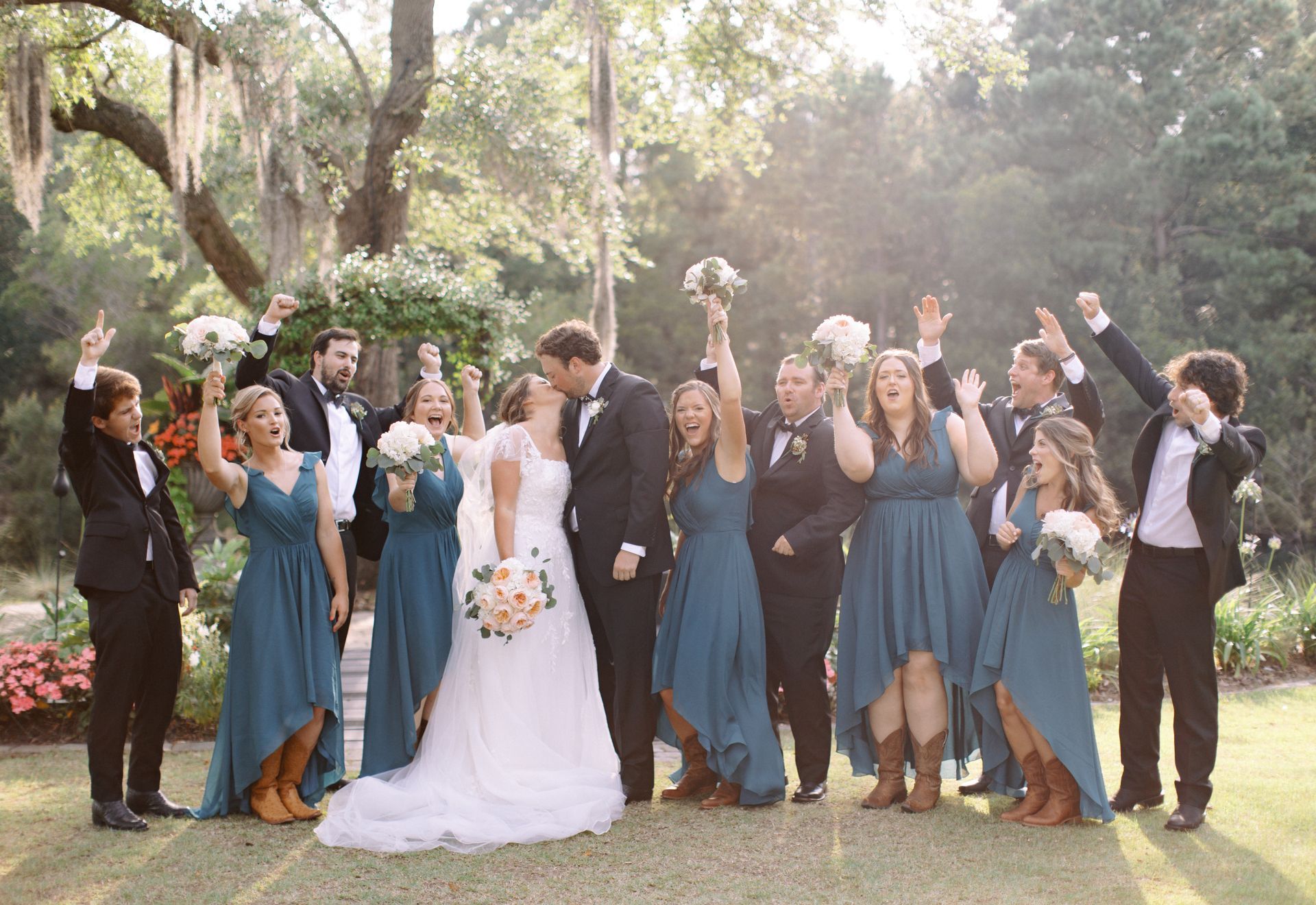 Wedding party celebrating outdoors in a sunlit garden, with the bride and groom in the center and guests cheering.