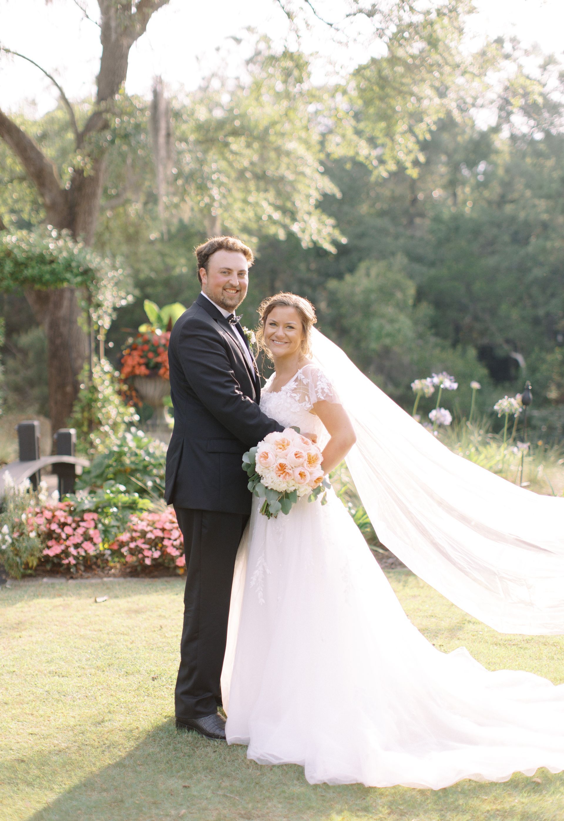 Bride and groom posing outdoors in a garden, holding a bouquet and smiling in sunlight