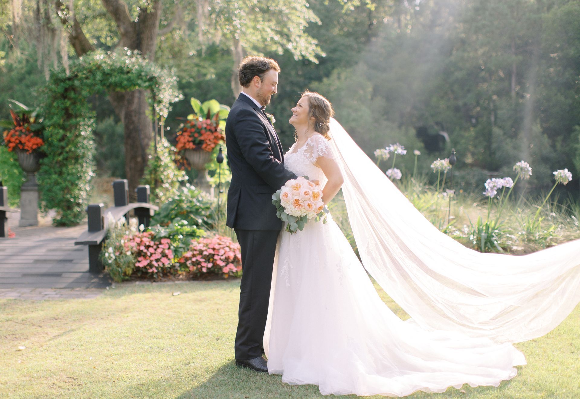 Bride and groom standing in a garden, smiling and holding hands beside a flower arch.
