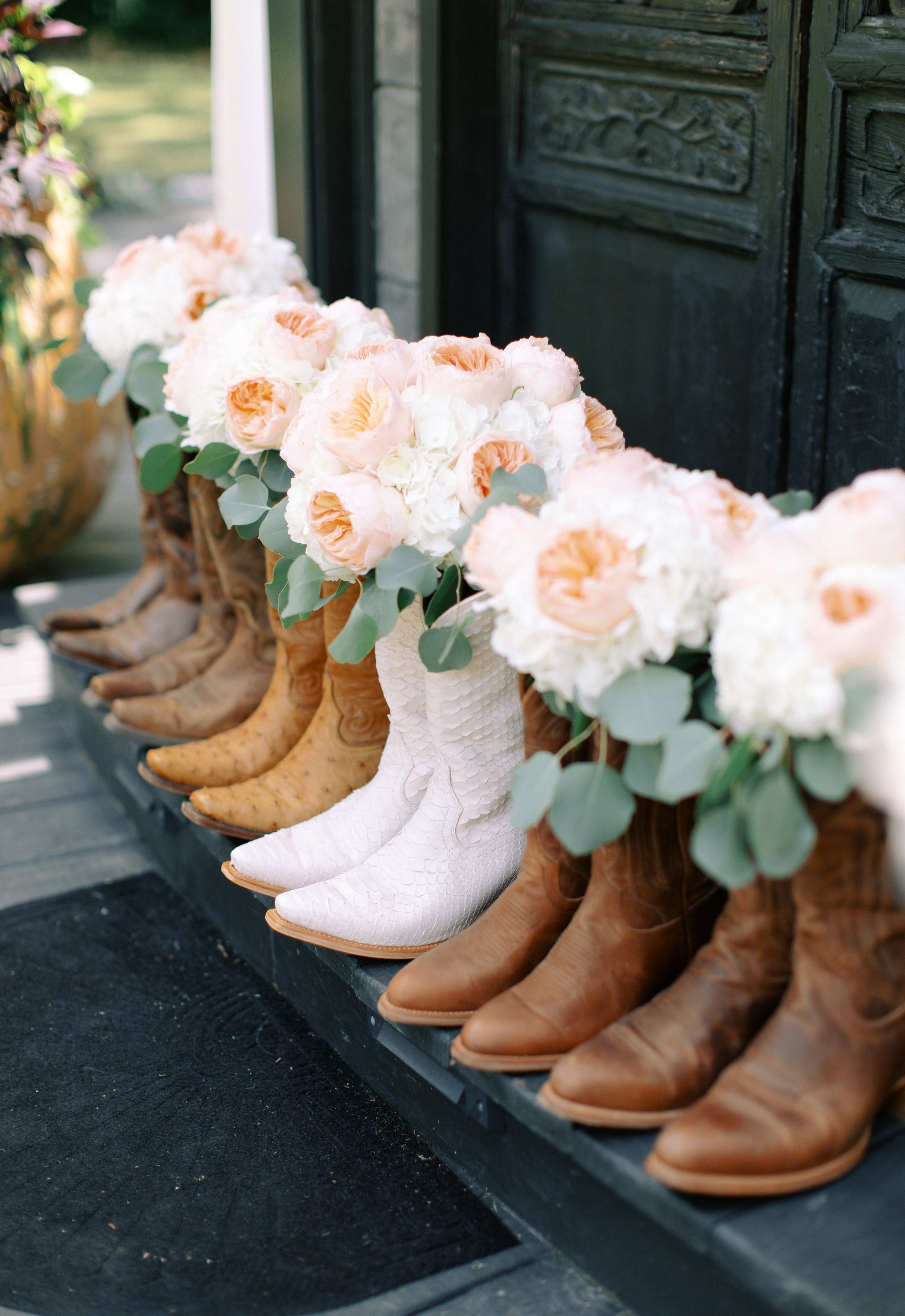 Row of rustic cowboy boots topped with white and peach flowers beside a dark doorway