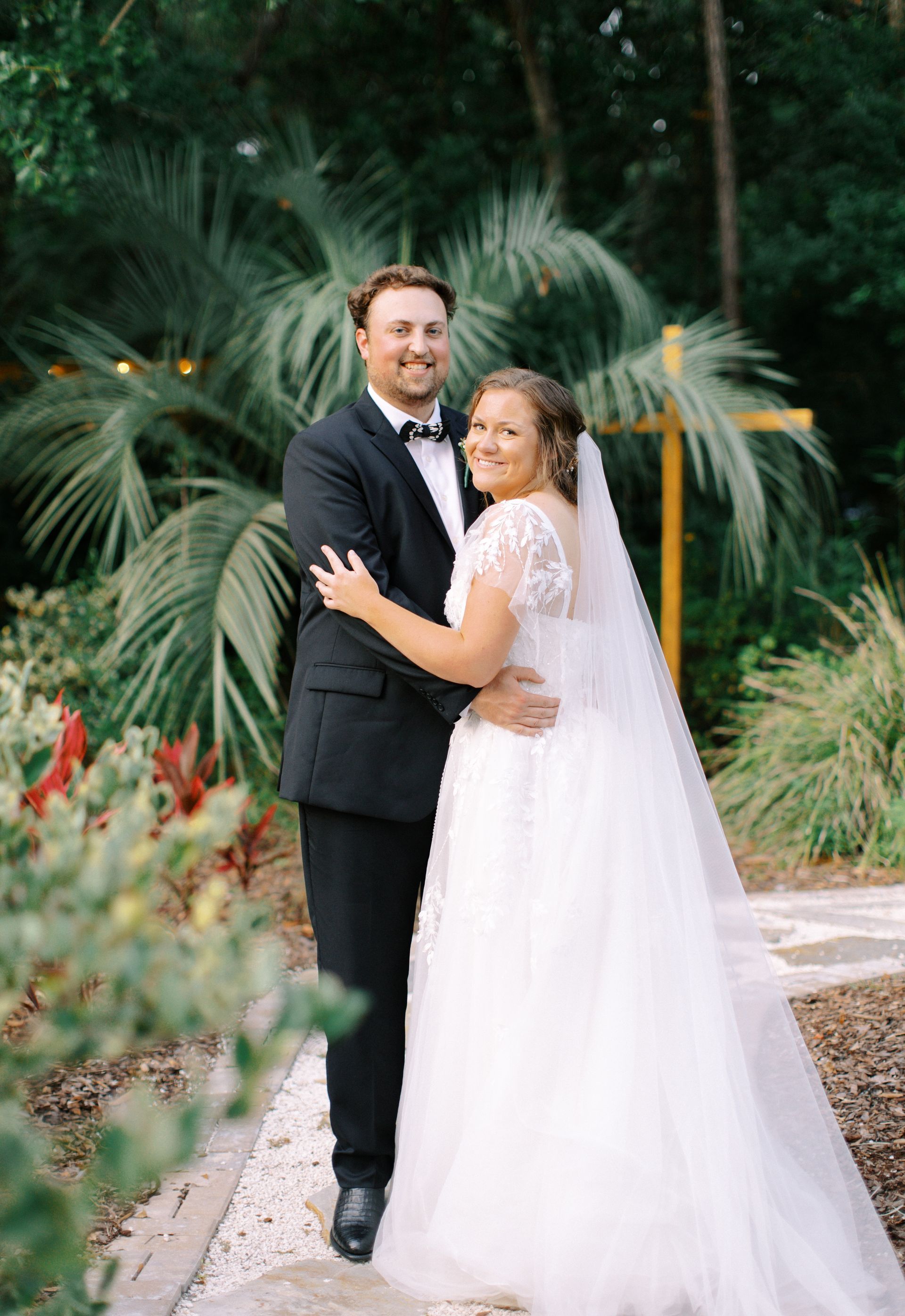 Couple in formal attire posing in a lush garden, the bride in a white veil and the groom in a black suit.