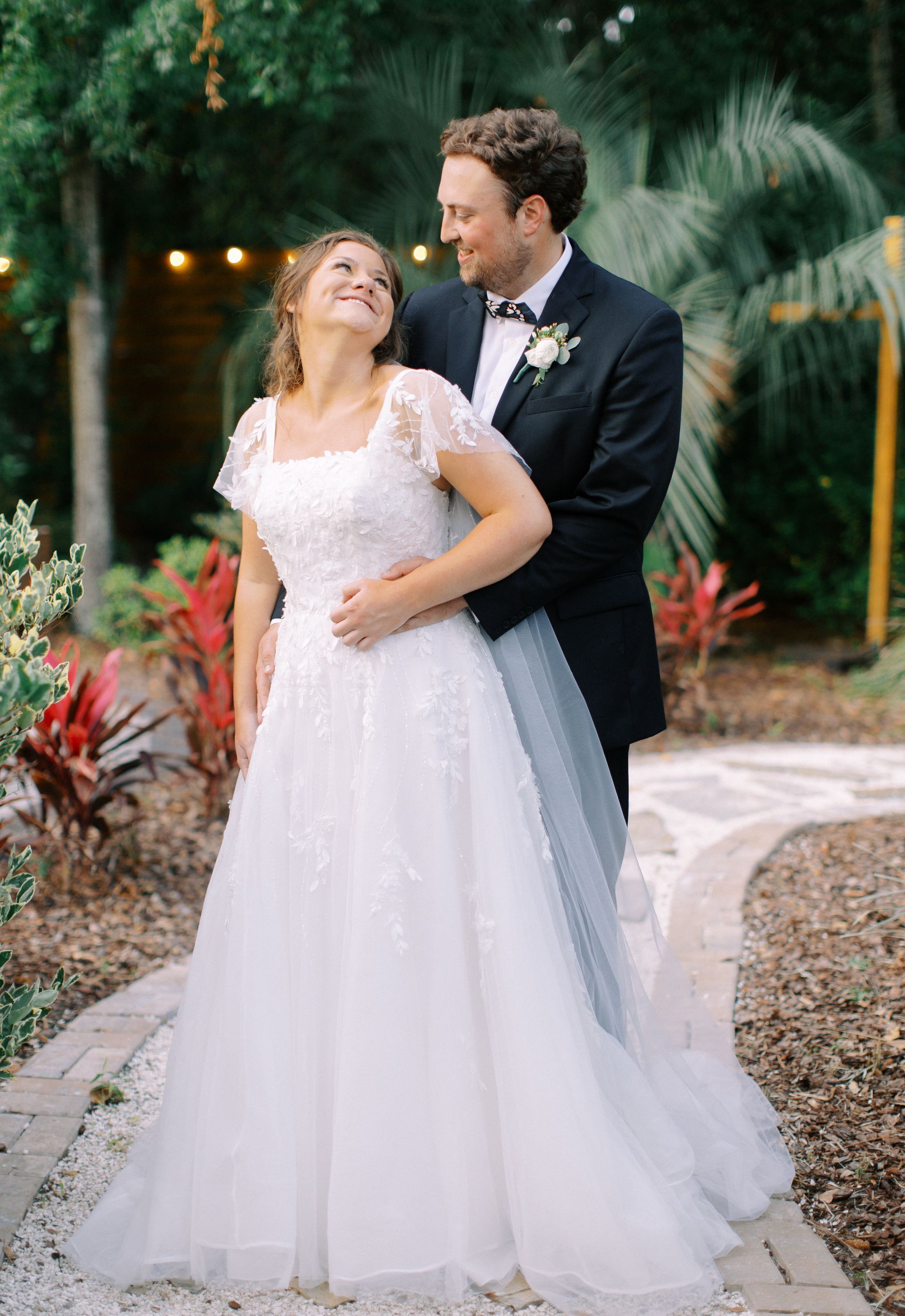 Bride and groom in wedding attire standing together outdoors, smiling and embracing amid greenery