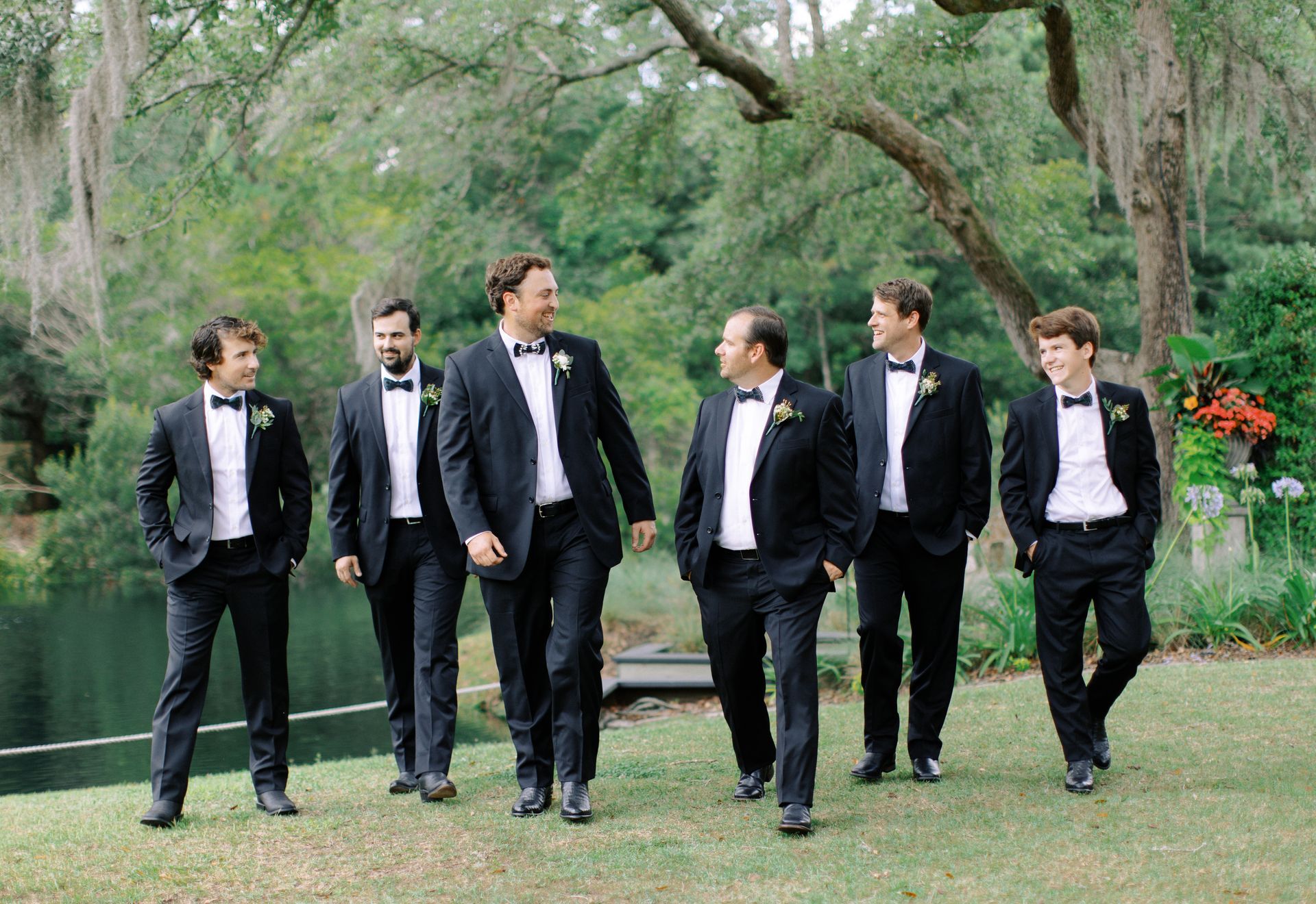 Six men in tuxedos walking together in a green garden with trees and flowers.