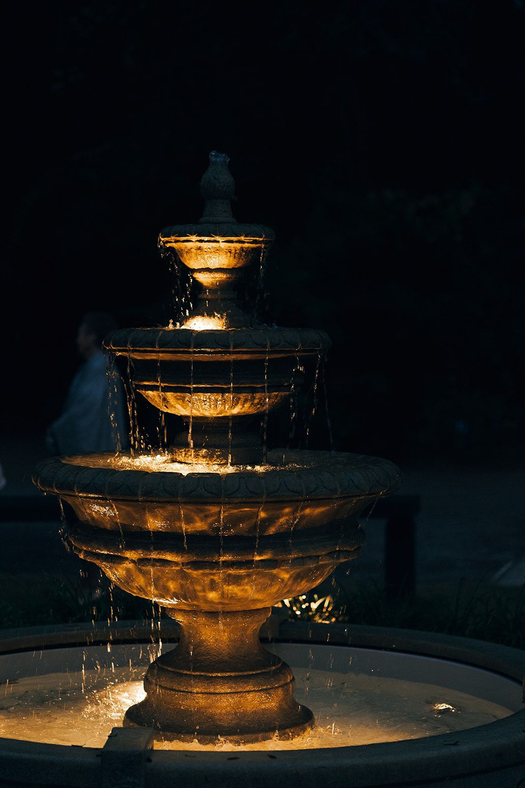 Lit tiered fountain at night, with warm golden lights glowing on the water and dark background