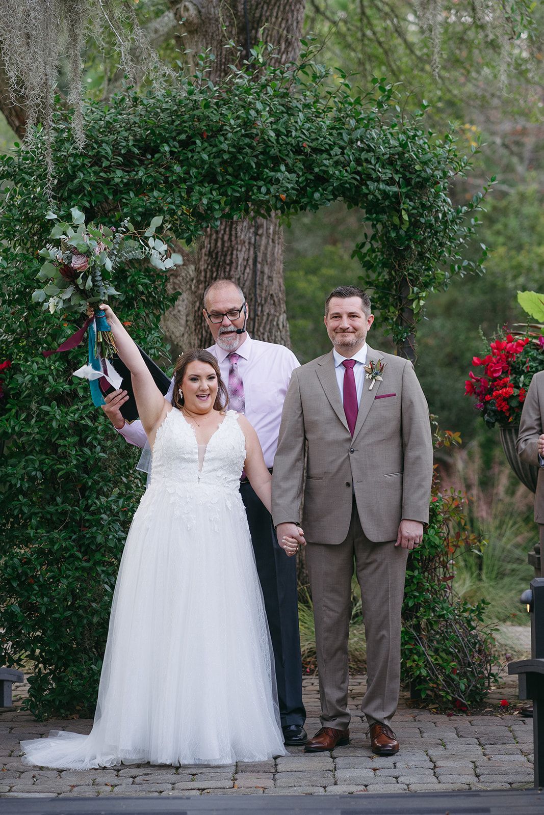 Wedding couple standing under a flower-covered arch, bride holding bouquet, officiant behind them, outdoors.