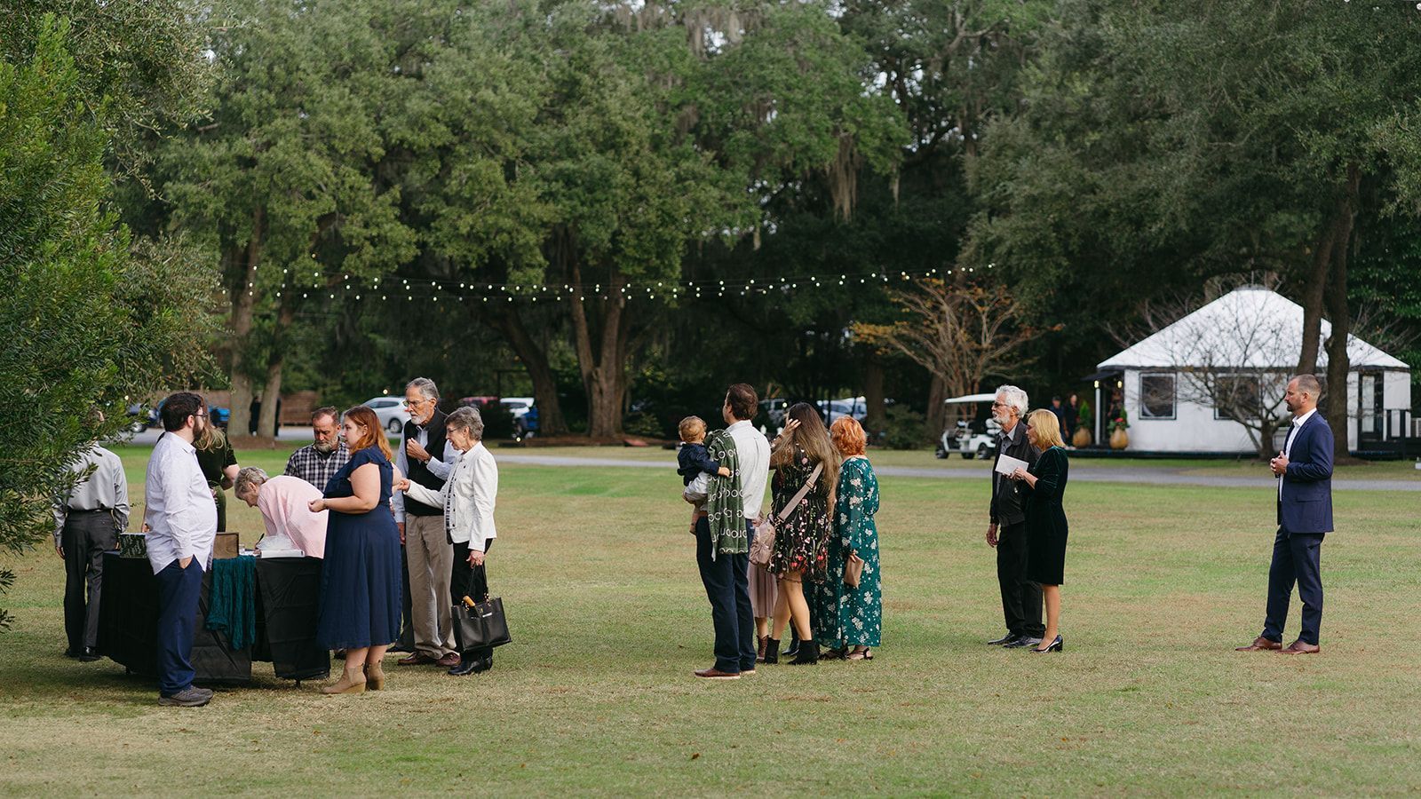 People gathered in small groups on a grassy field near a white tent, outdoors in a park.
