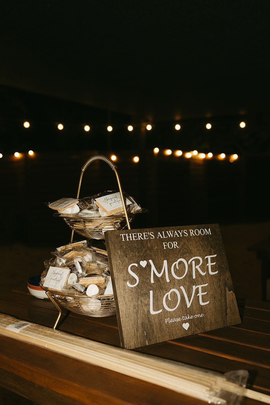 S'more Love sign beside a basket and s'mores display on a dimly lit table with string lights in the background