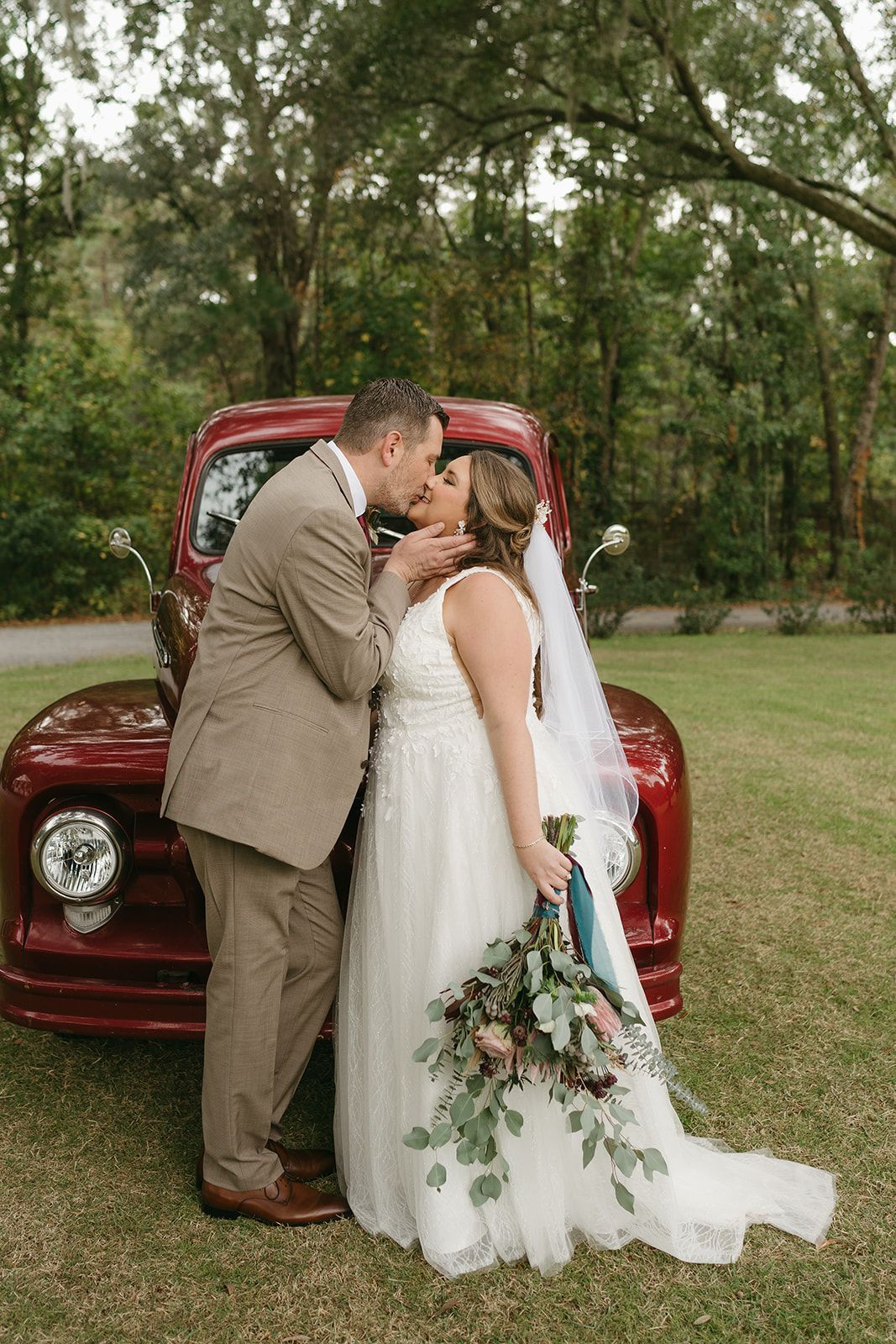 Bride and groom kissing in front of a red vintage car outdoors