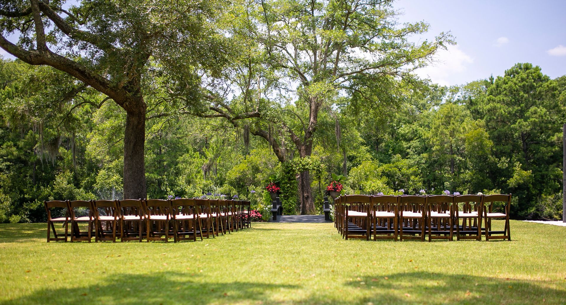 Outdoor wedding setup with wooden chairs on a grassy lawn beneath trees and a pink aisle backdrop