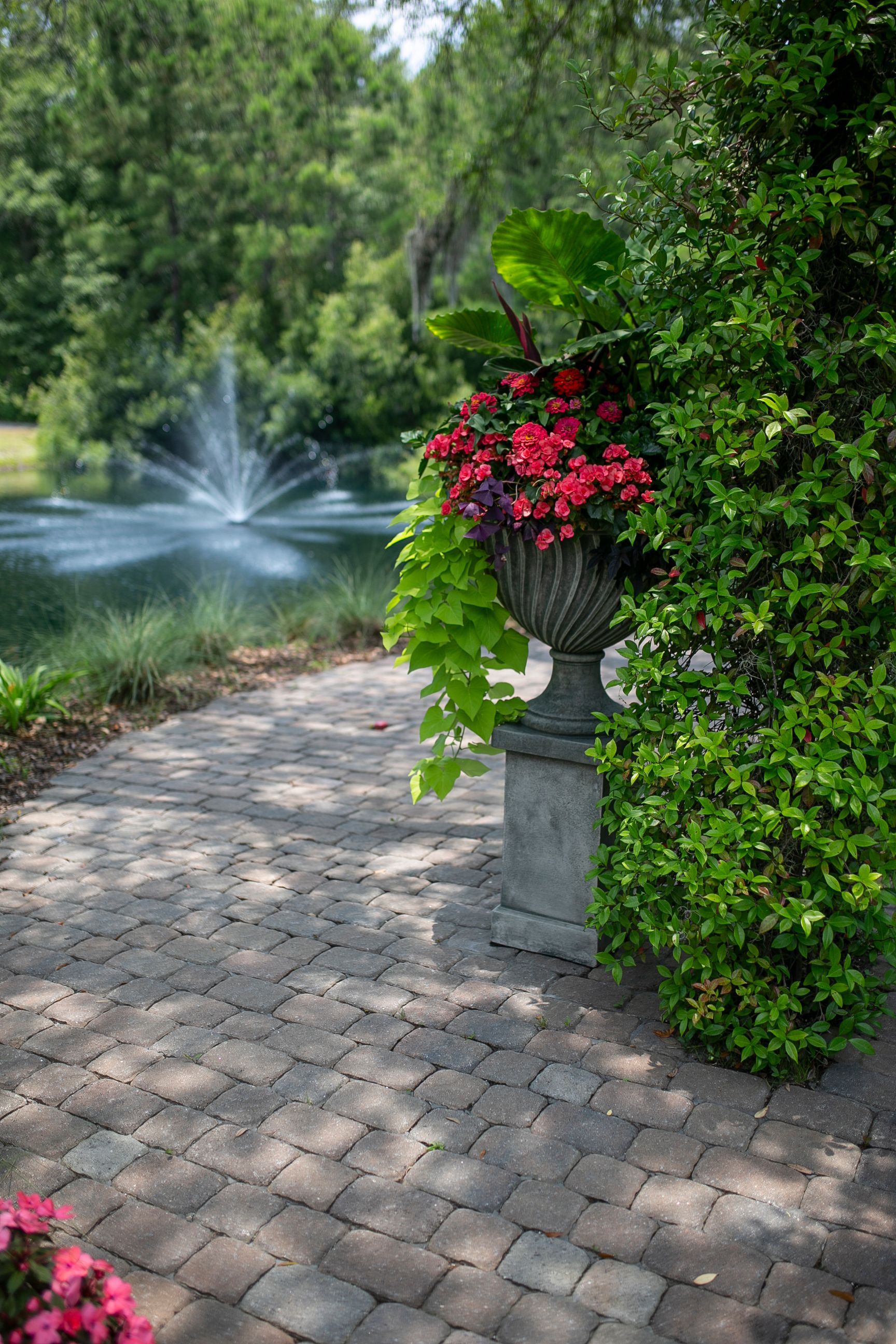 Brick garden path beside a pond fountain, with a red flower basket on a stone pedestal and green foliage