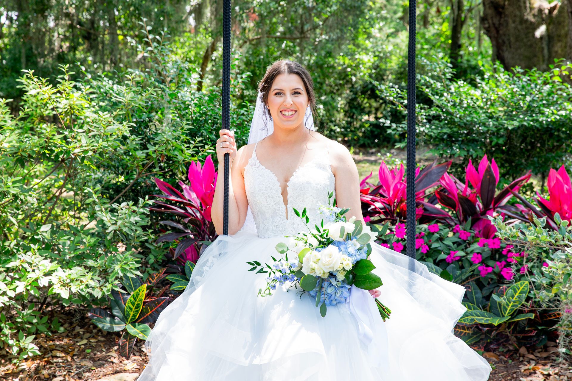 Bride in a white gown holding a bouquet, seated among bright pink flowers in a lush garden.