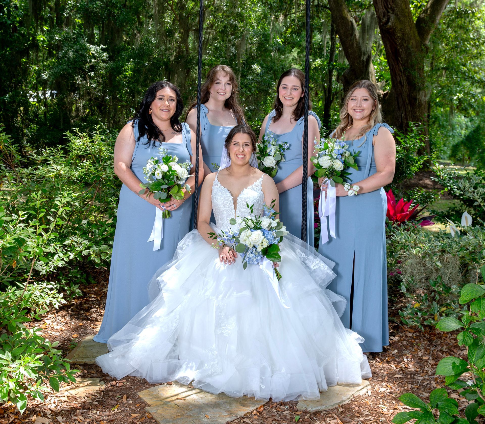 Bride seated with four bridesmaids holding bouquets in a lush garden setting