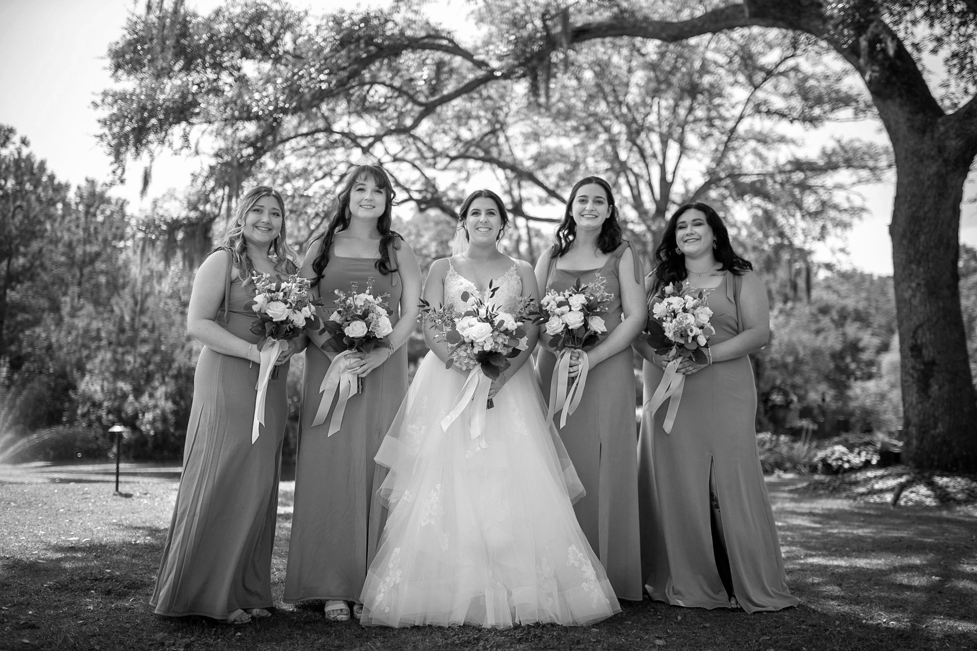 Five women in formal dresses pose outdoors, holding bouquets beneath a large tree.