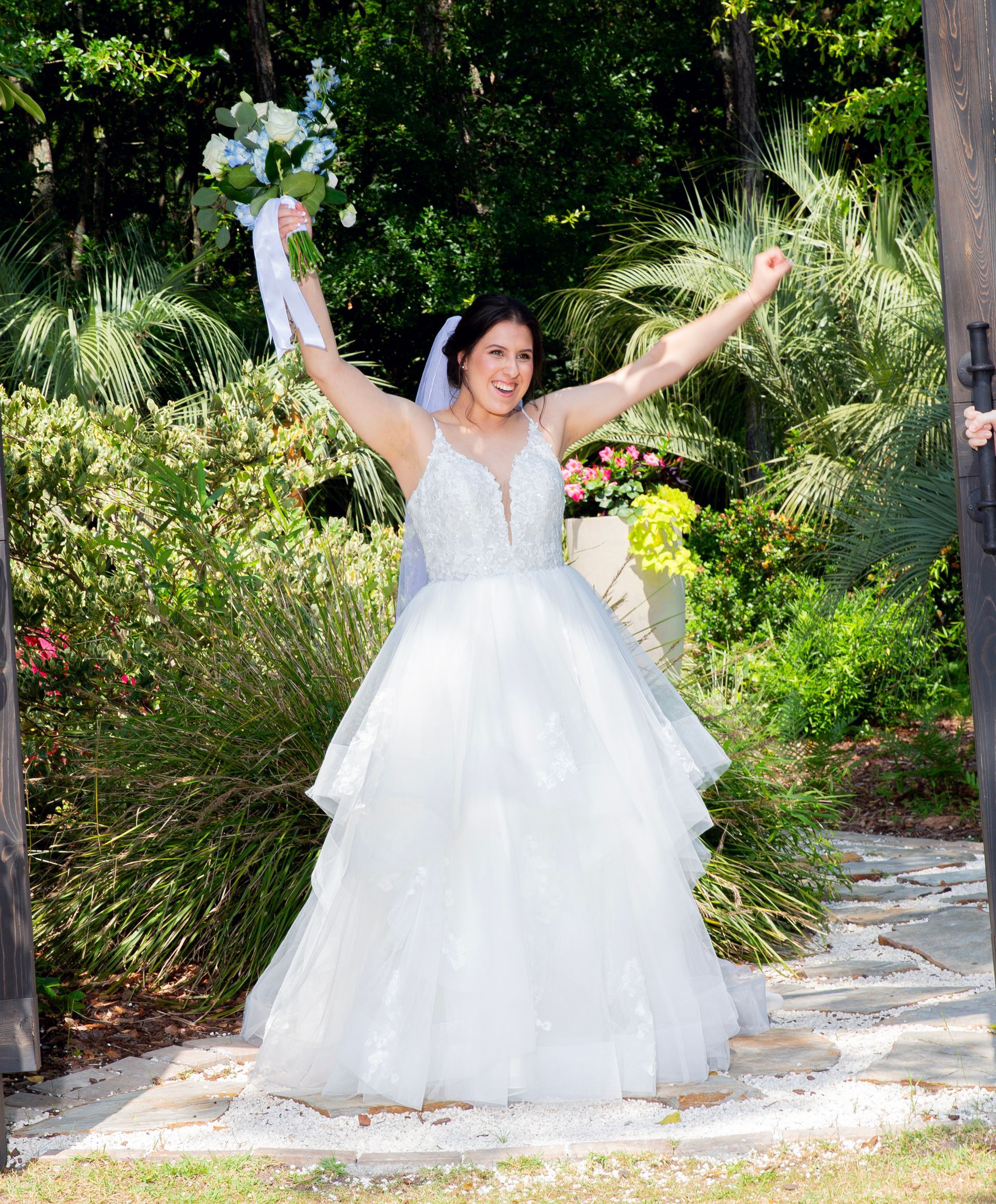 Bride in a white wedding dress with arms raised, holding a bouquet in a lush garden path.