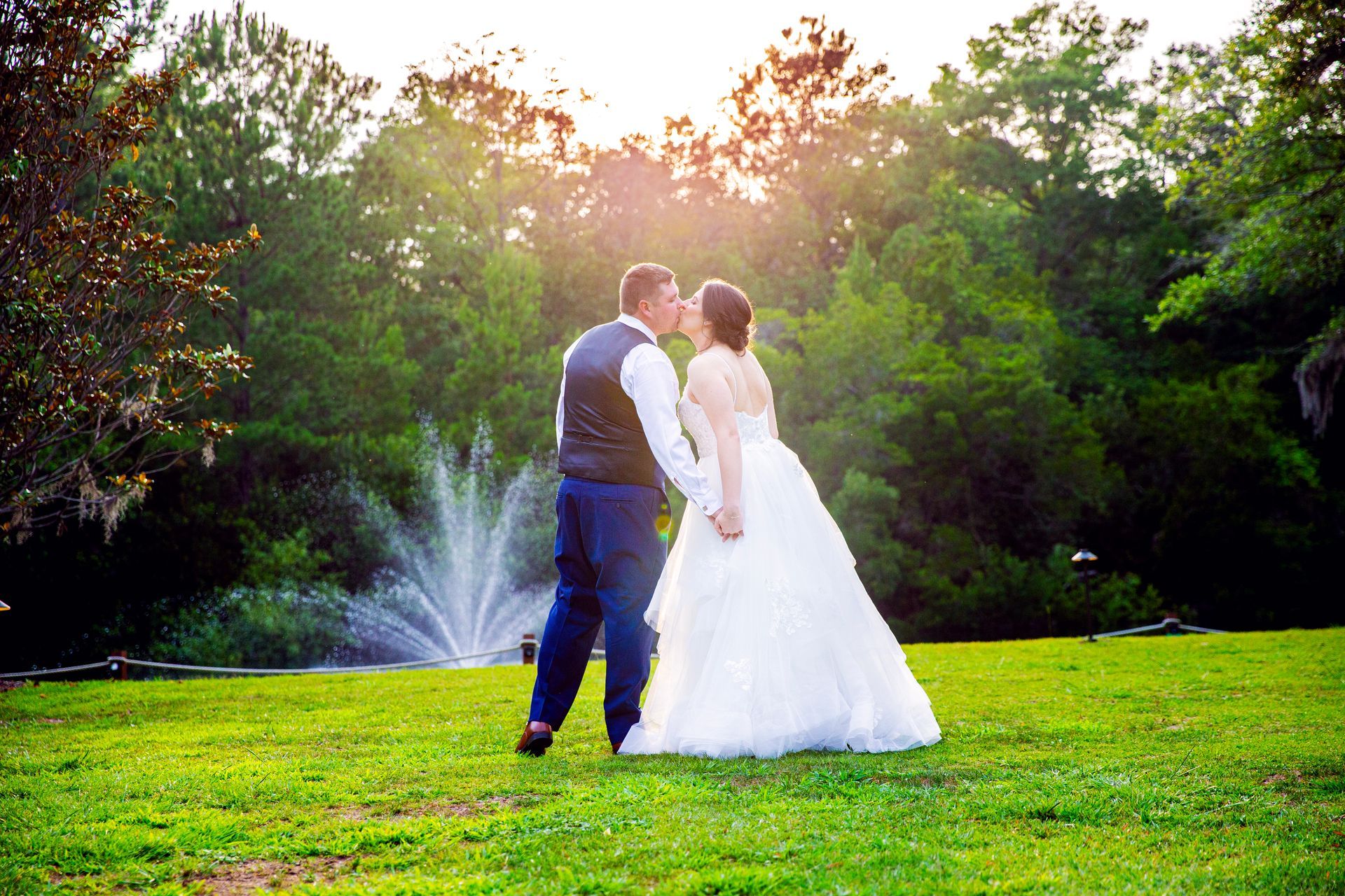 Bride and groom kissing on a sunlit lawn with a fountain in the background