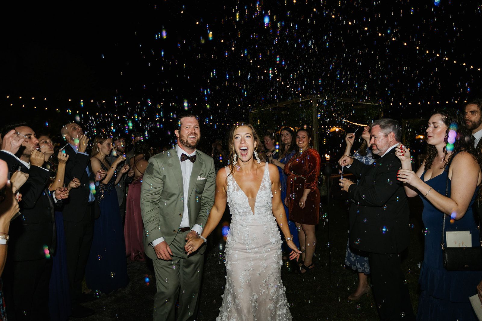 Newlyweds walking through confetti at a nighttime wedding reception, guests applauding nearby