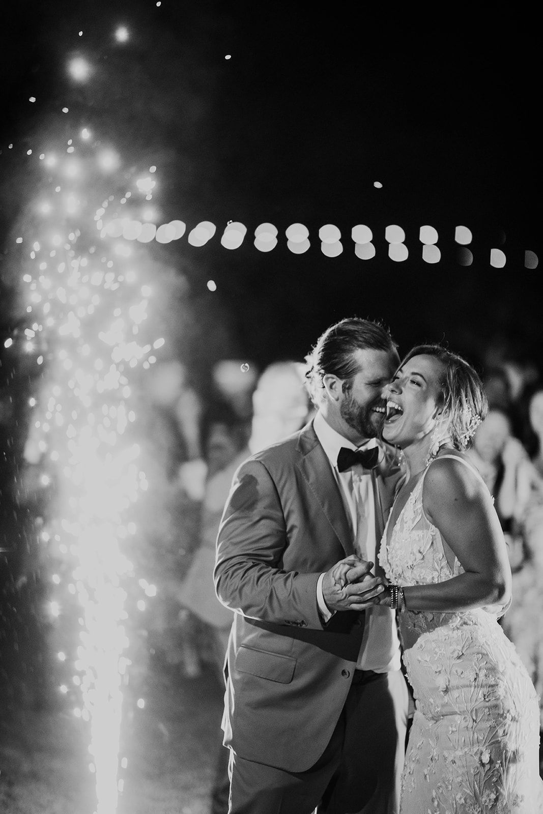 Newlyweds dancing at a nighttime wedding, smiling under string lights and sparklers