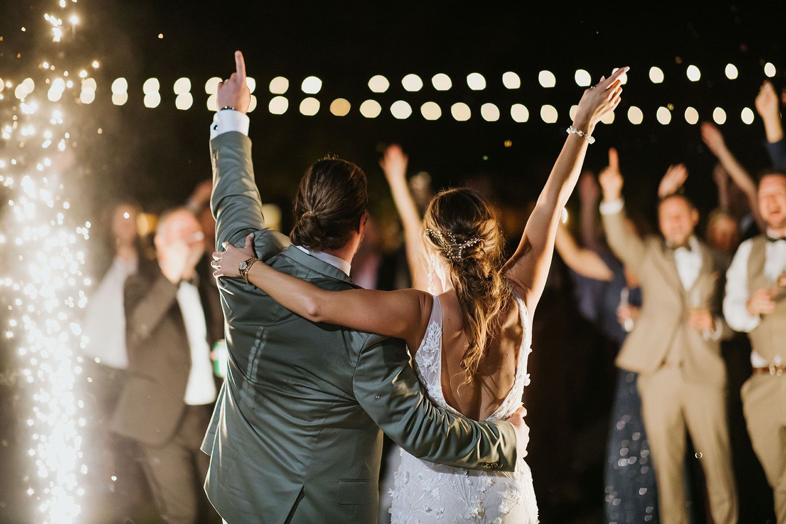 Newlyweds dancing with arms raised at a nighttime celebration, surrounded by cheering guests and string lights