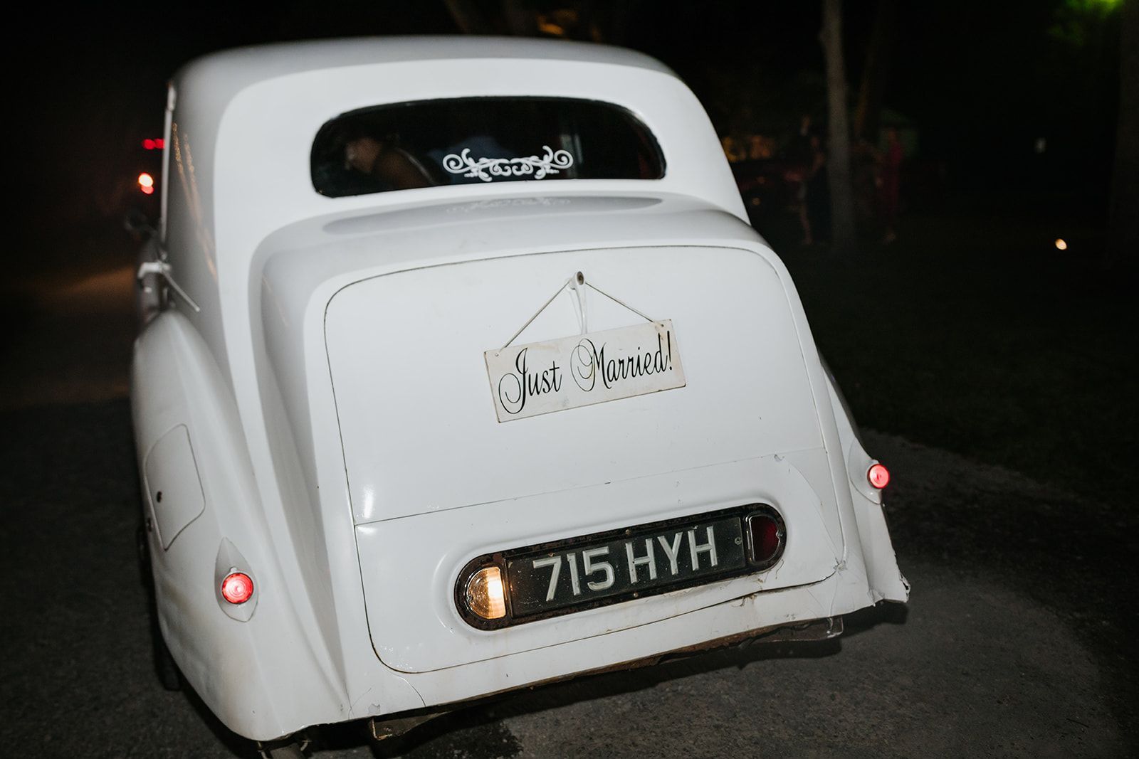 White vintage car viewed from the rear at night, with illuminated license plate and small rear window decals