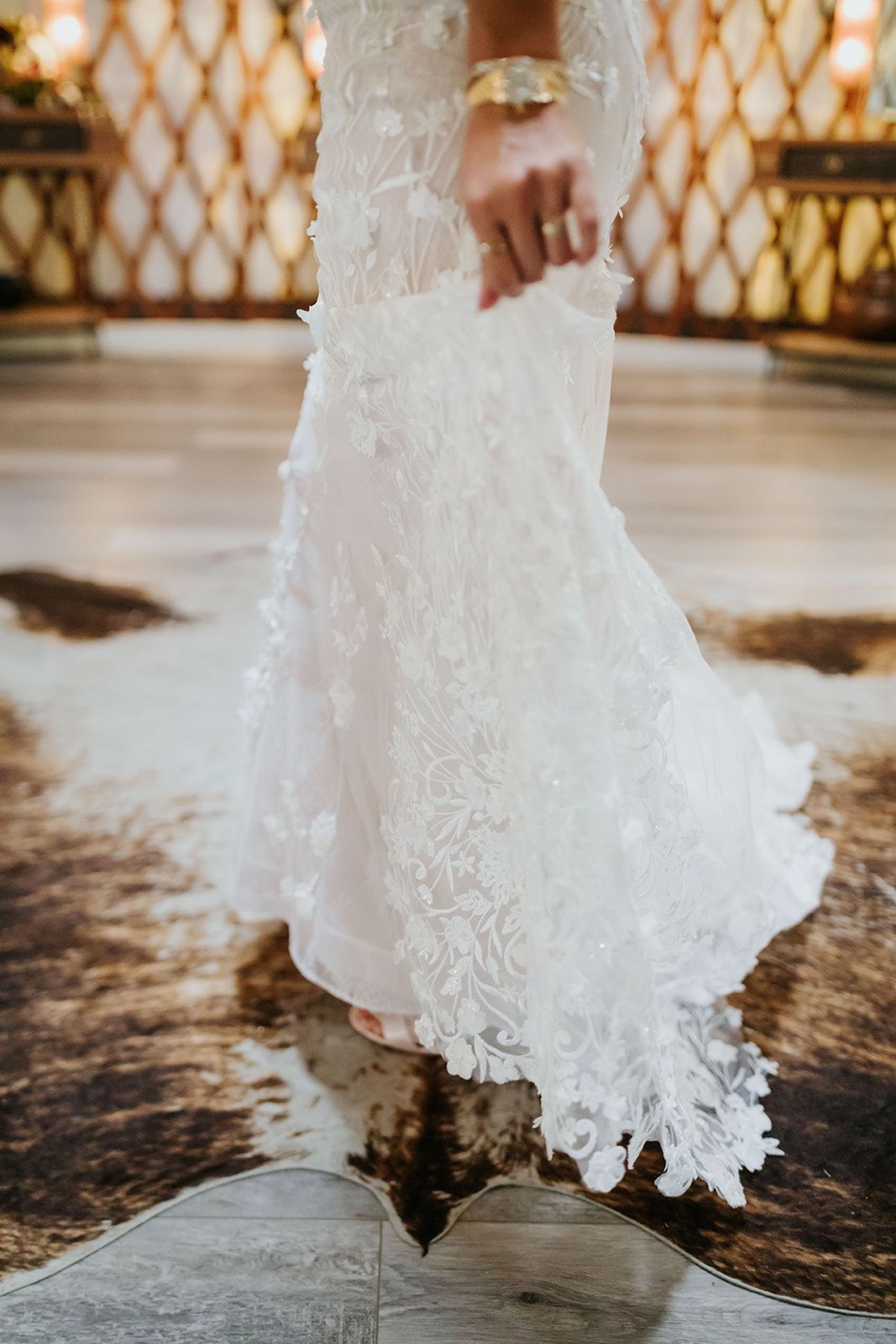 White lace wedding dress hem with a hand at the waist on a patterned rug in a warm-lit interior