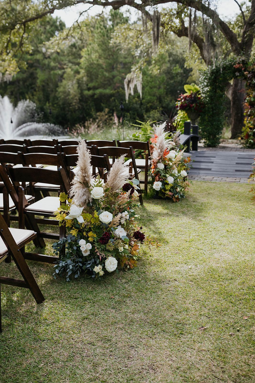 Outdoor wedding aisle with white floral arrangements, wooden chairs, and a fountain in a garden setting