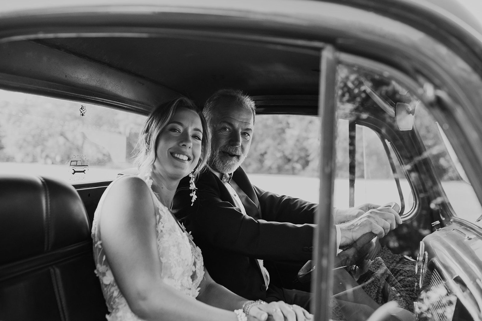 Couple smiling in a vintage car, seated side by side with the driver holding the wheel.
