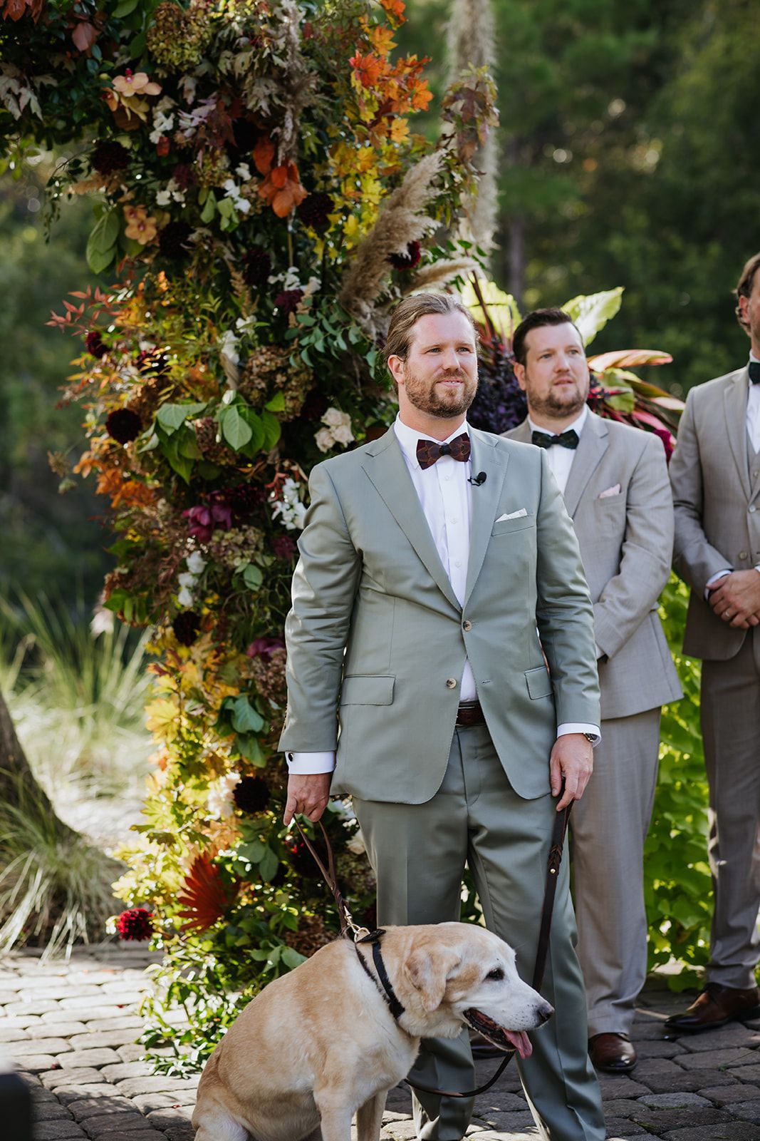 Man in a light gray suit standing beside a tan dog at an outdoor floral wedding ceremony