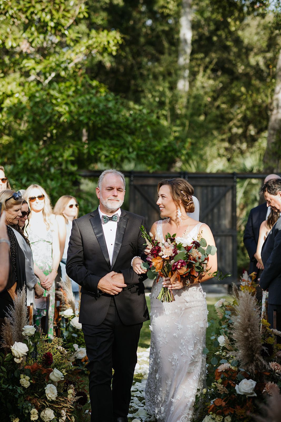 Bride in a white gown holding a bouquet, walking with an older man outdoors at a garden wedding ceremony