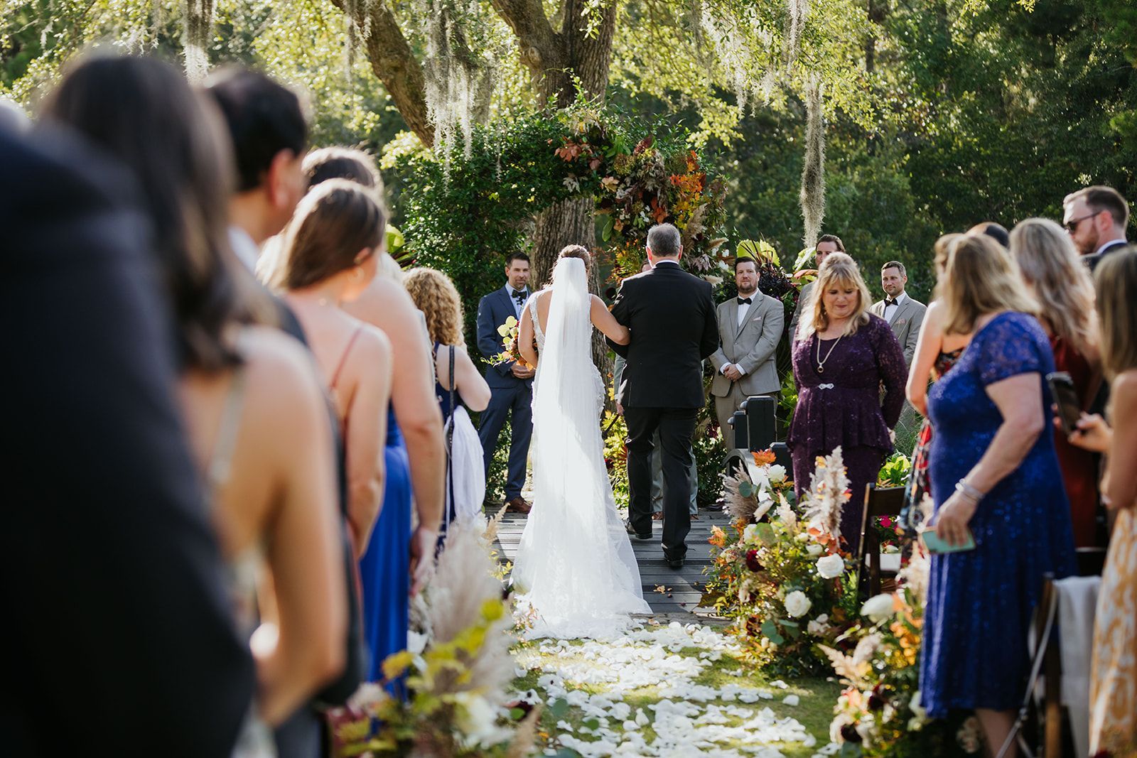 Wedding ceremony outdoors with bride and groom standing under trees, guests lining the aisle