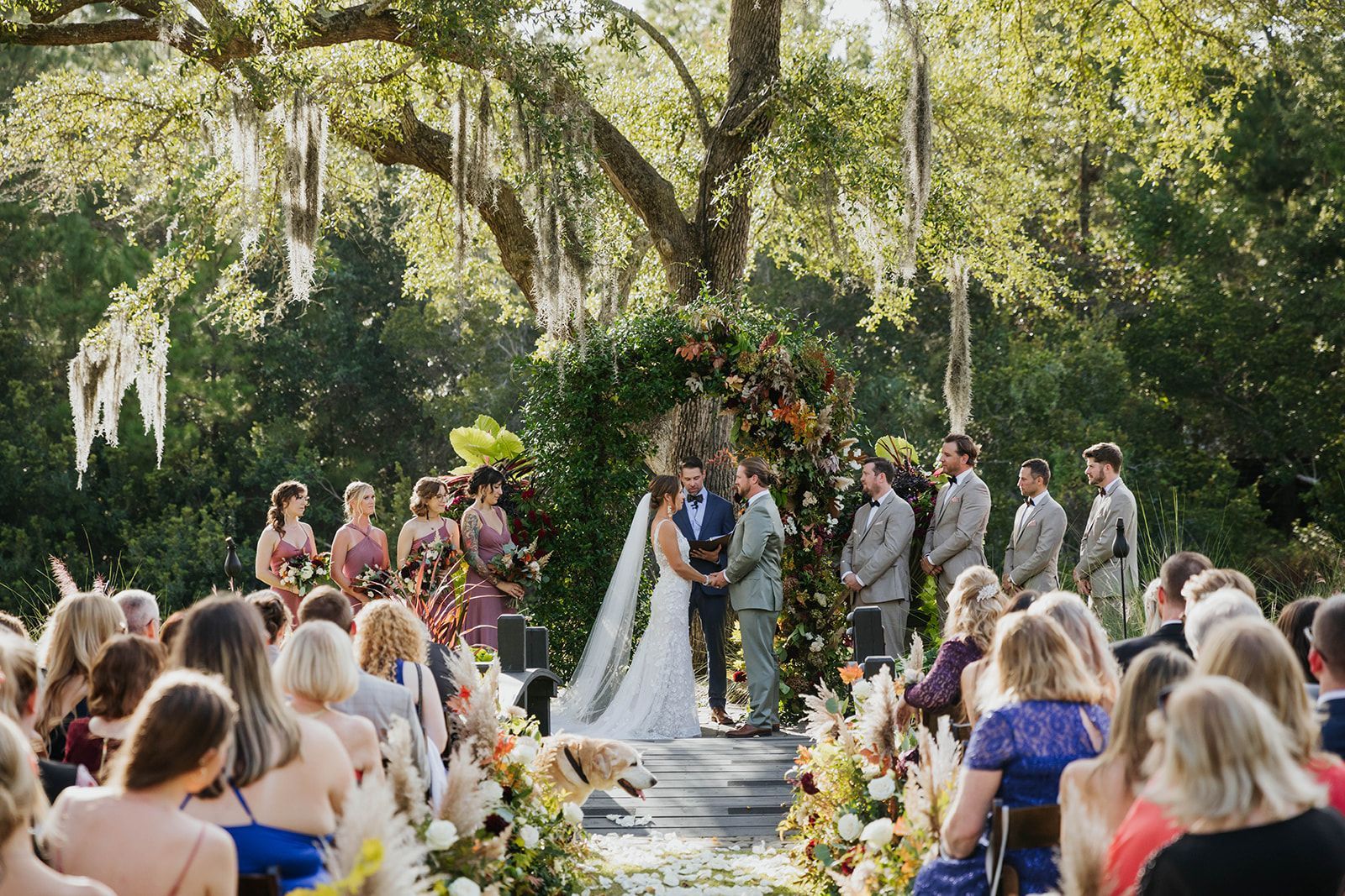 Outdoor wedding ceremony under trees with bride, groom, and guests seated on both sides of the aisle