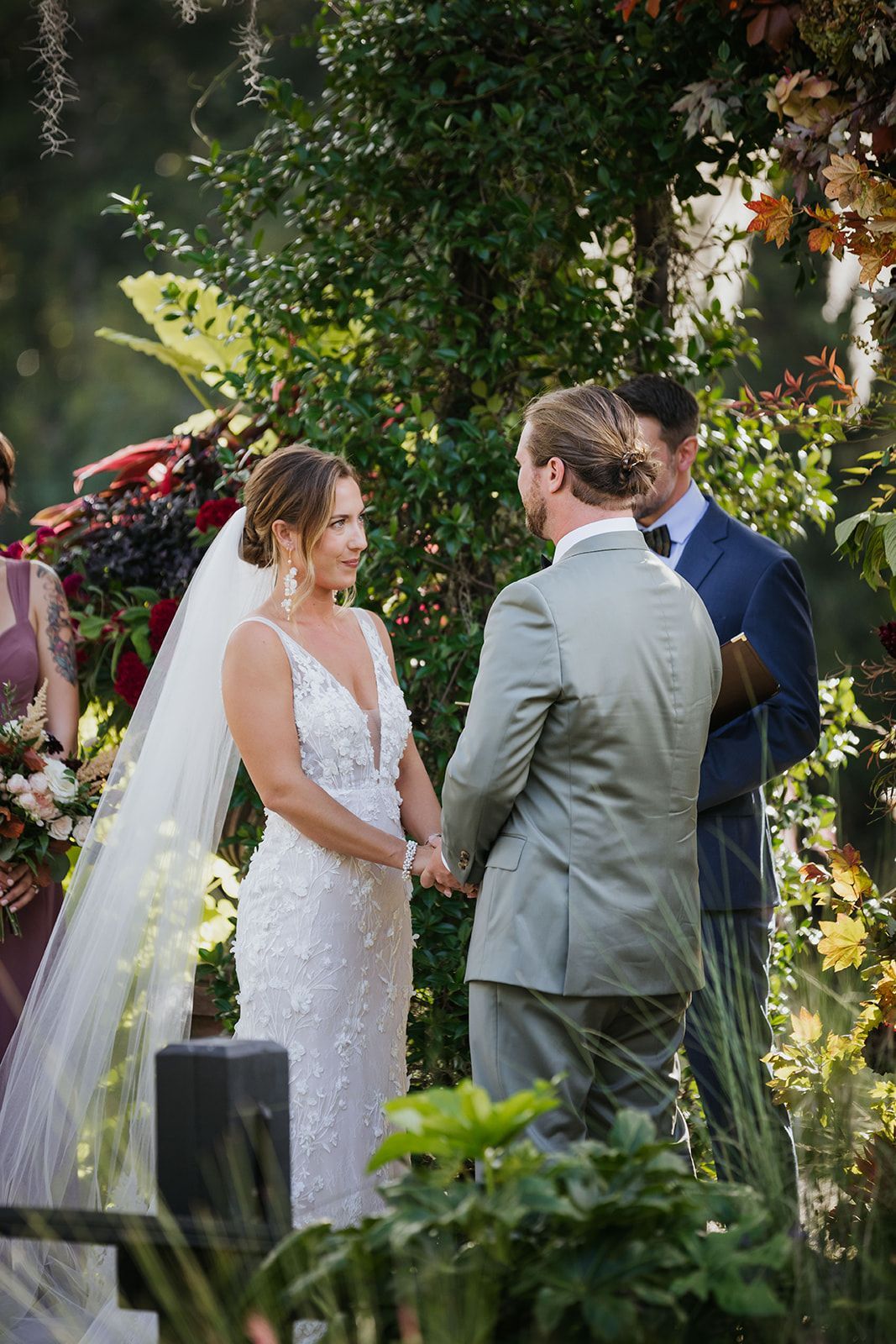 Bride and groom exchanging vows outdoors under greenery, with an officiant nearby