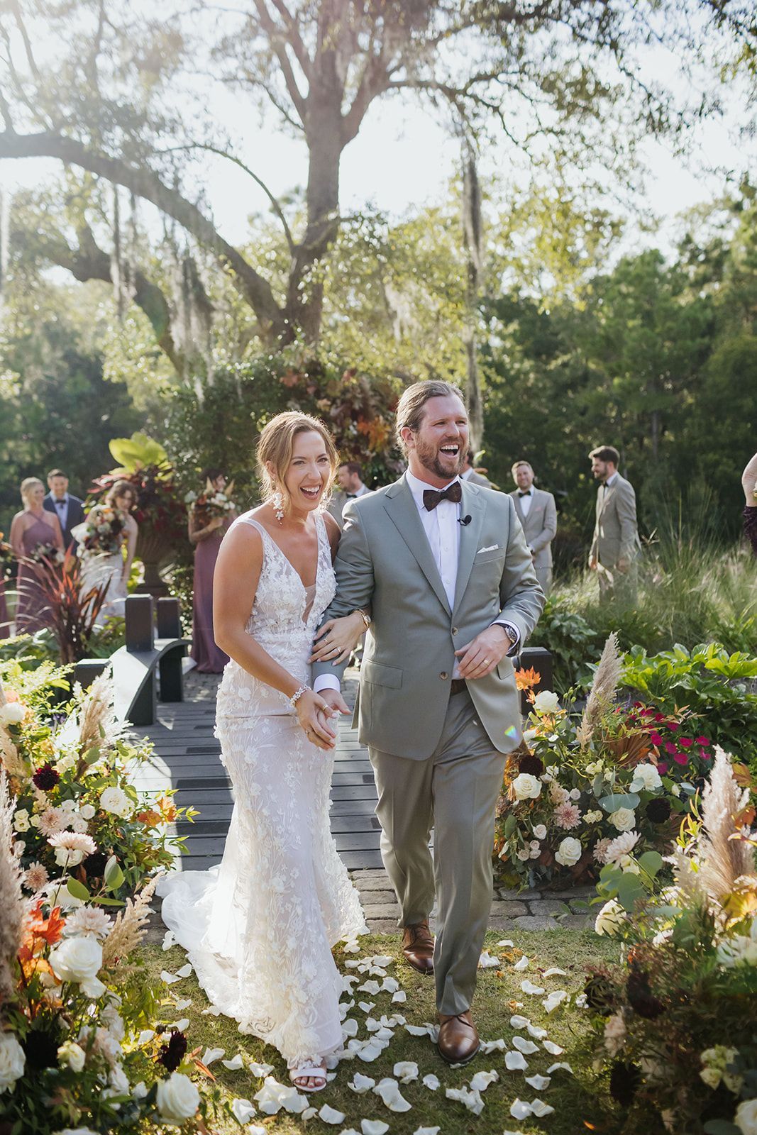 Bride and groom walk down a flower-lined outdoor aisle, smiling in a sunlit garden.