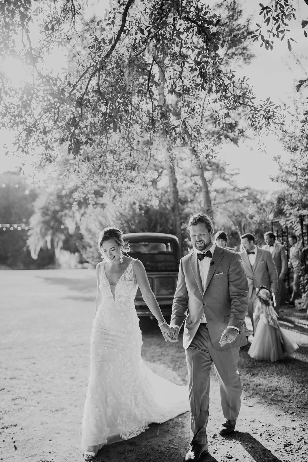 Couple holding hands in wedding attire outdoors under trees, with guests in the background.