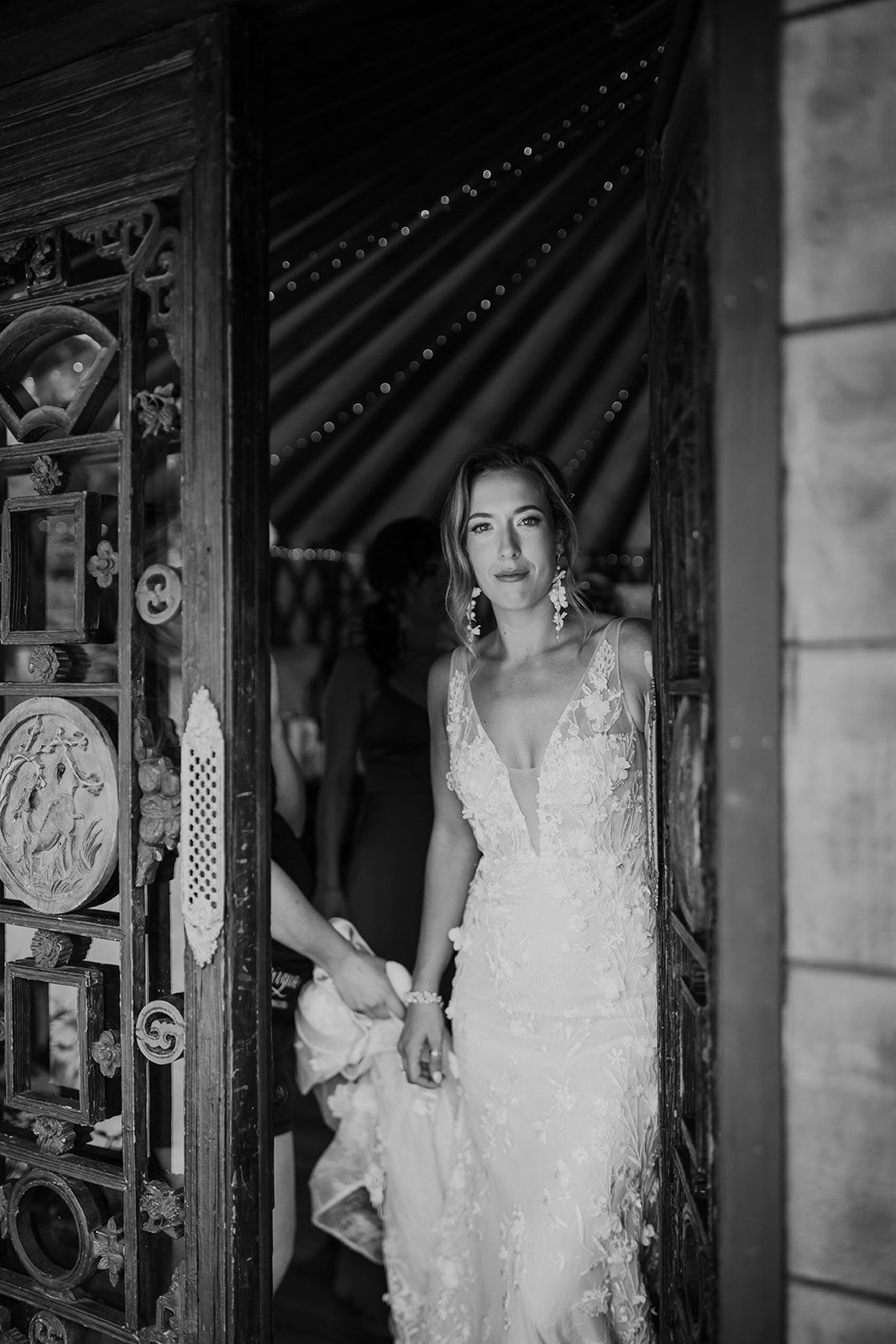 Woman in a white lace dress standing in a doorway, holding hands with someone off-frame.