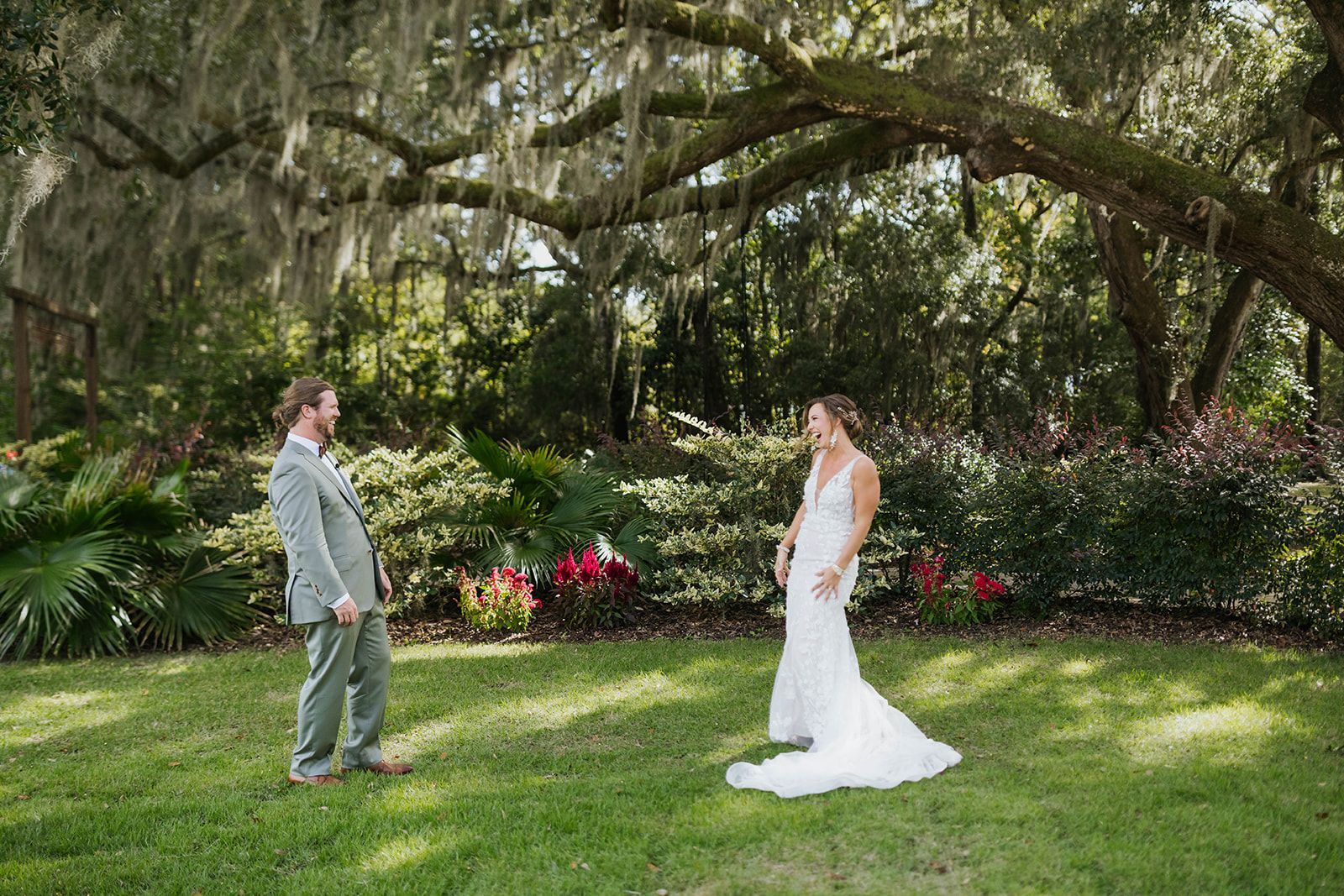 Two people face each other on a grassy lawn beneath a large tree, dressed for a wedding ceremony.