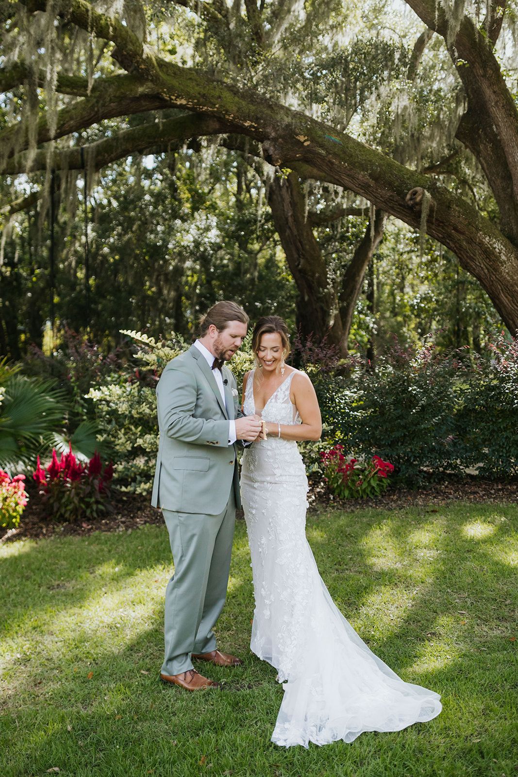Bride and groom in light wedding attire standing together in a sunlit garden under large oak trees