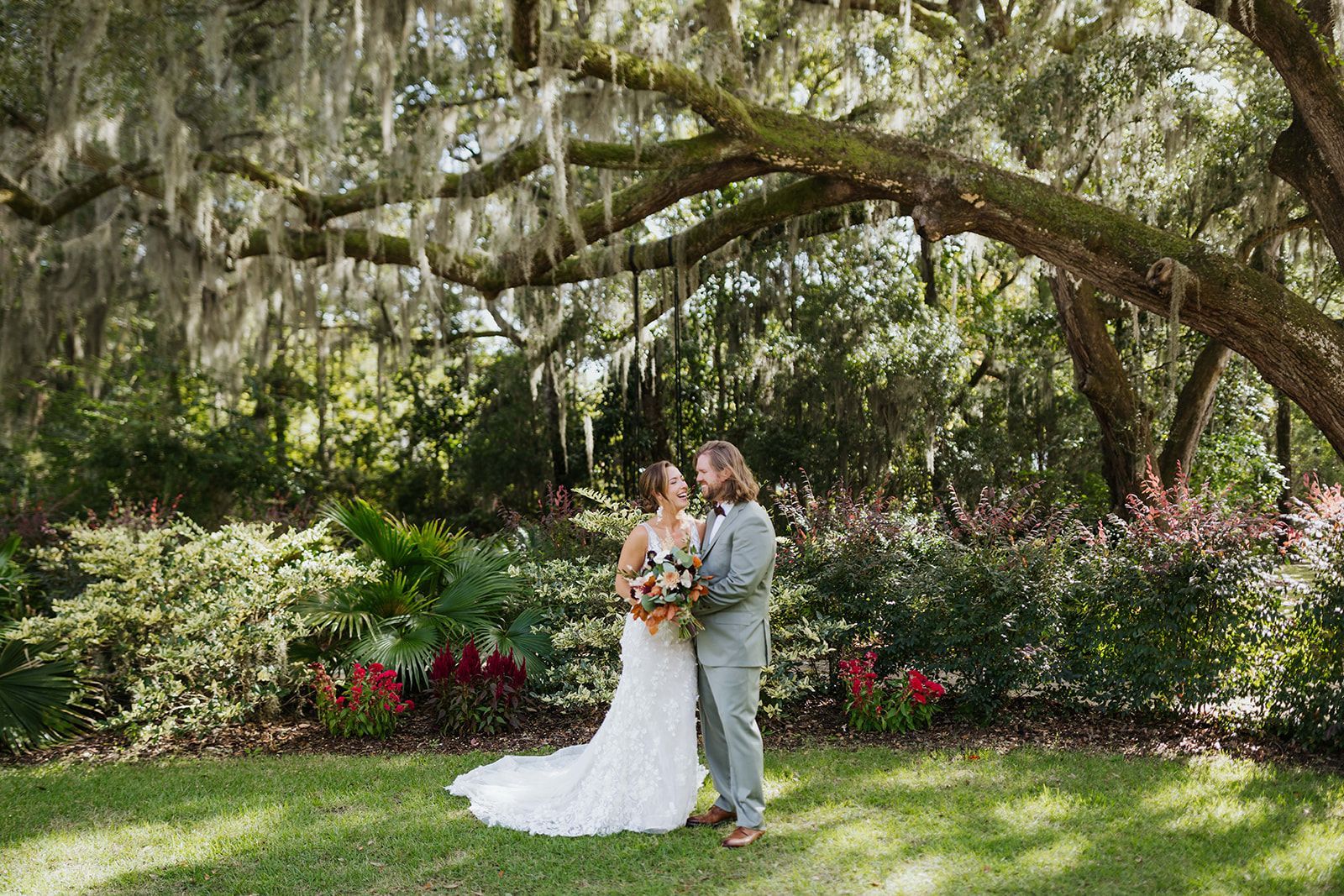 Couple in wedding attire standing in a lush garden under arching trees