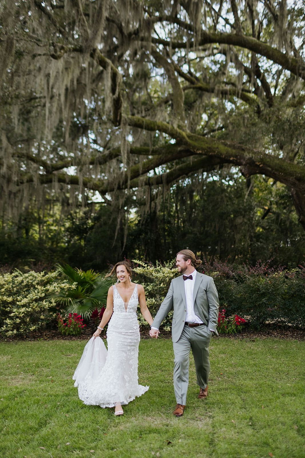 Newlyweds holding hands on a grassy garden path beneath a large moss-draped tree