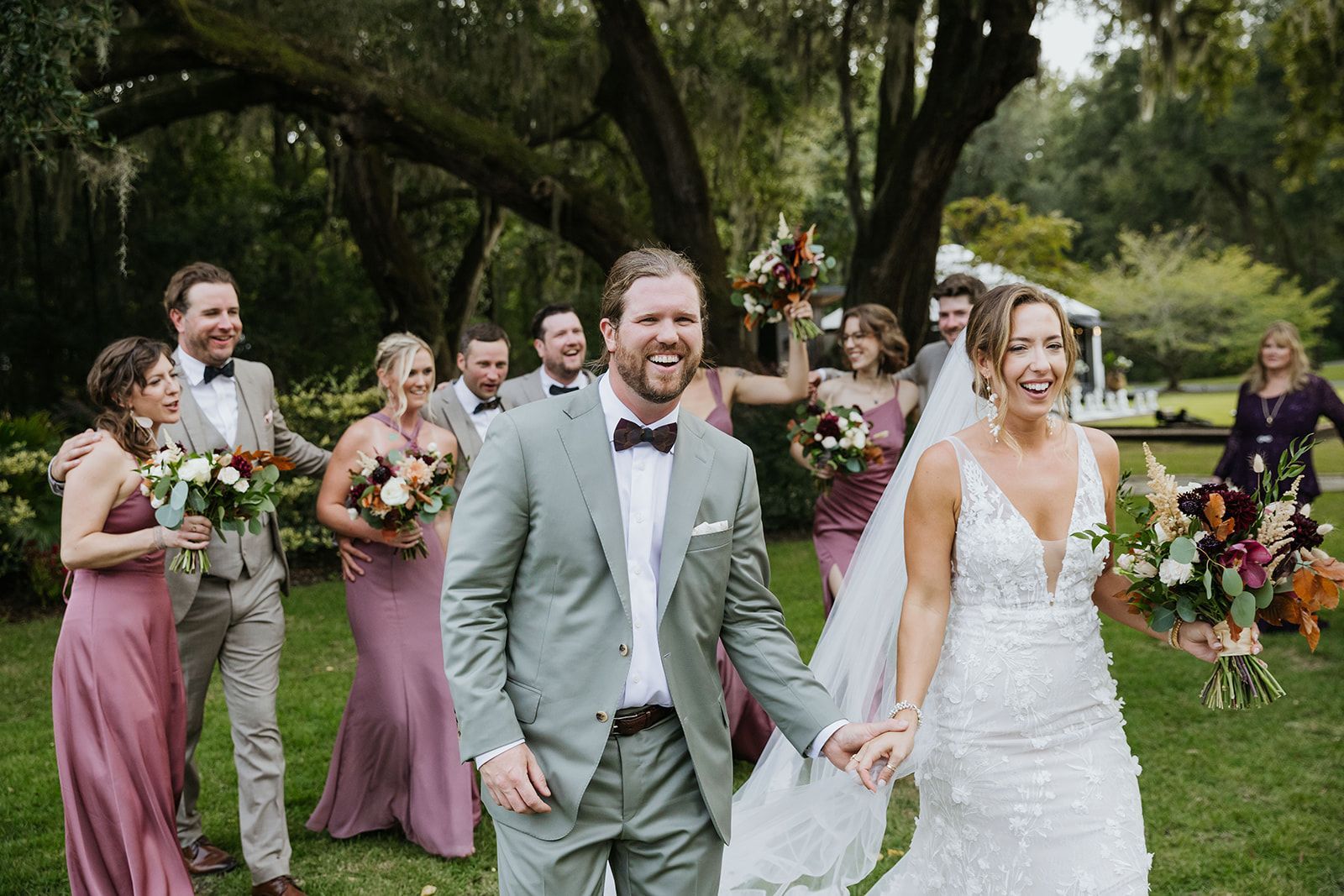 Wedding party outdoors with bride and groom walking hand in hand, bridesmaids in mauve dresses behind them.