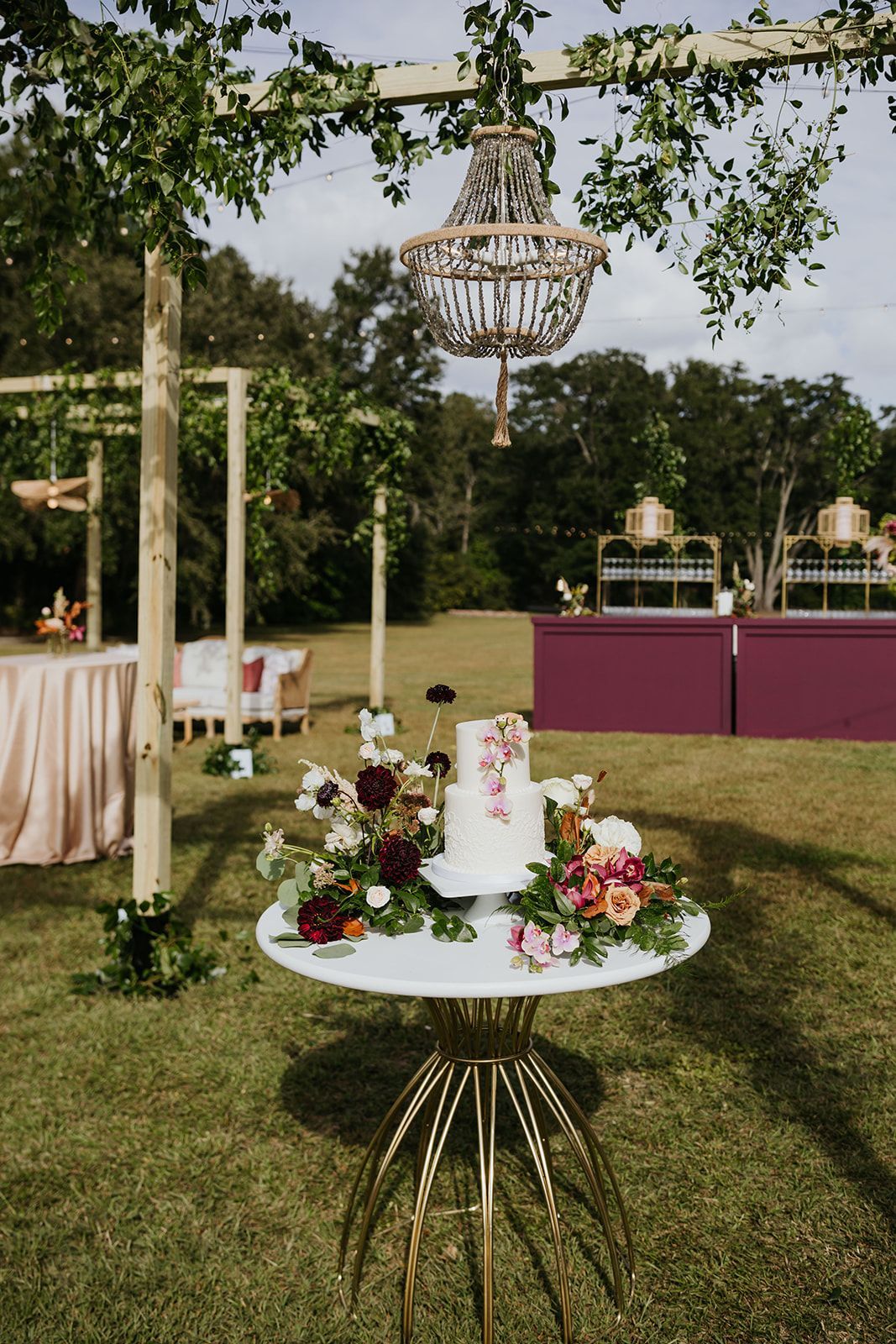 Outdoor wedding table with white cake, flowers, and hanging chandelier under a tree on a lawn