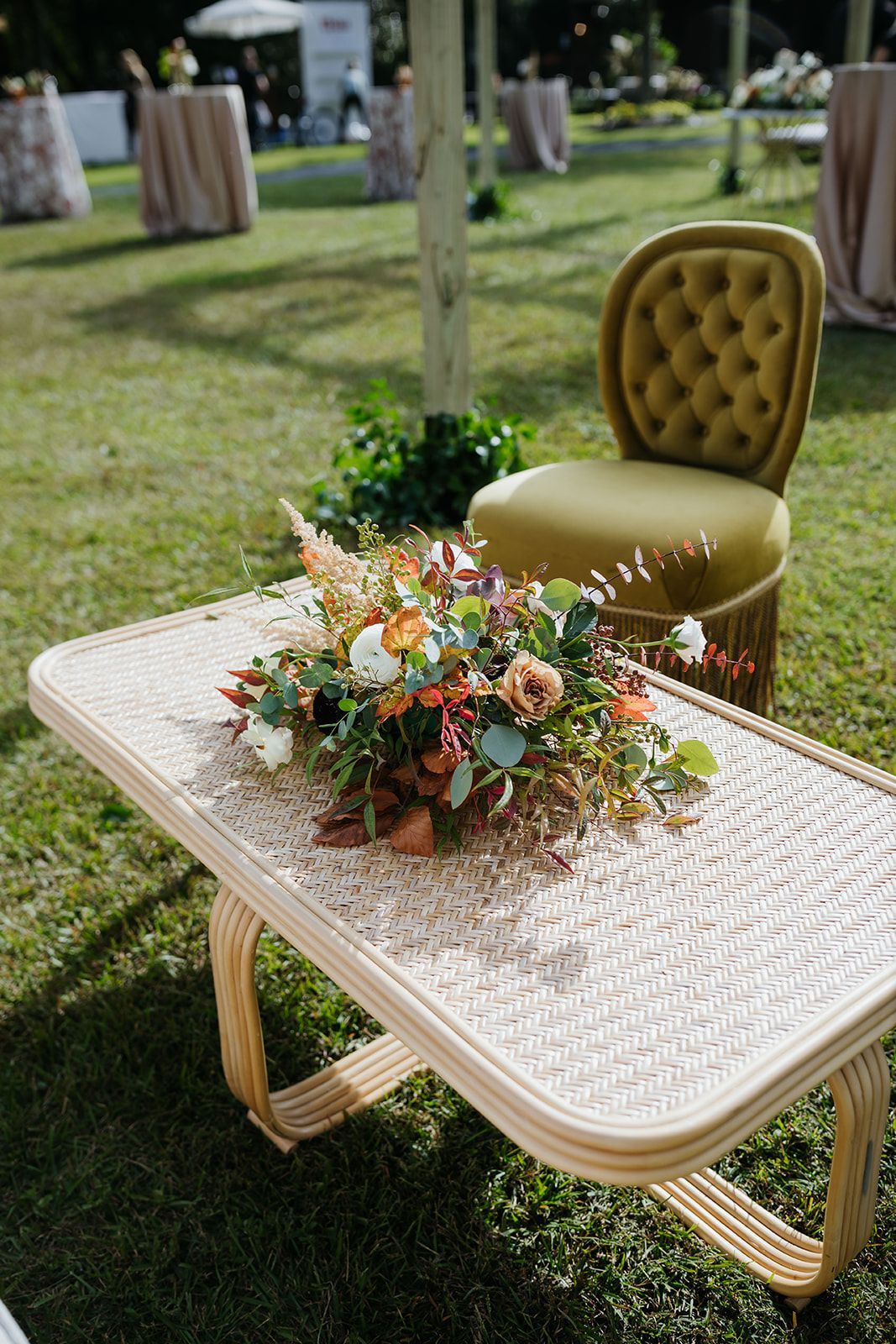 White woven table with pastel floral centerpiece on grass beside a mustard tufted chair outdoors