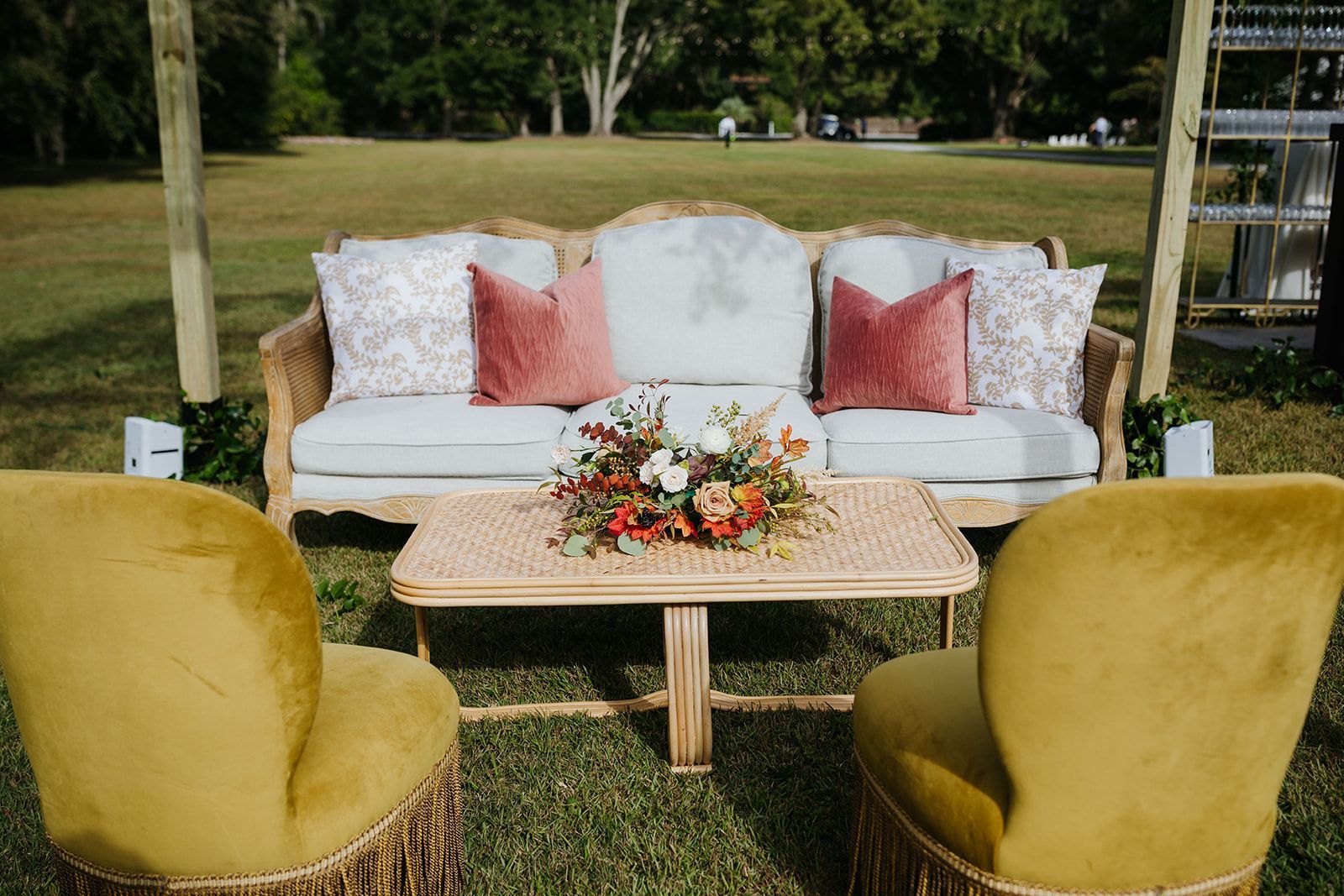 Outdoor lounge setup with white sofa, pink pillows, floral centerpiece, and two mustard chairs on a lawn