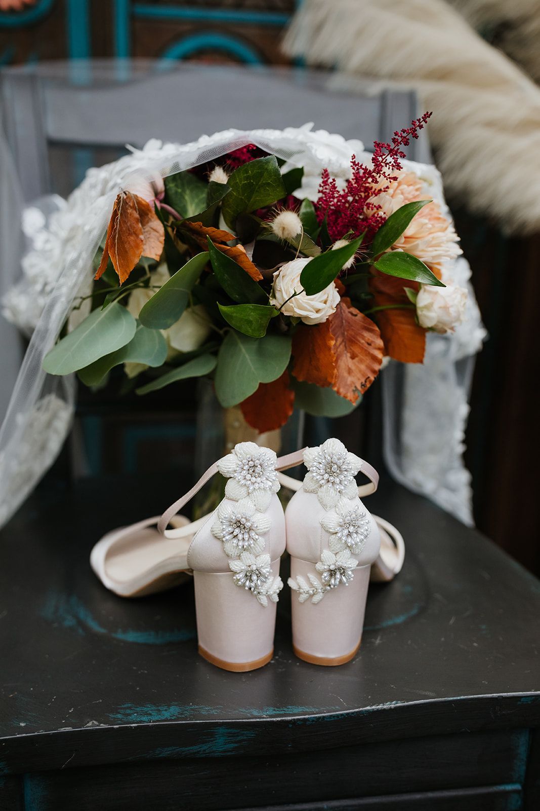 White lace bridal heels beside a bouquet with white, green, and burgundy flowers on a dark table