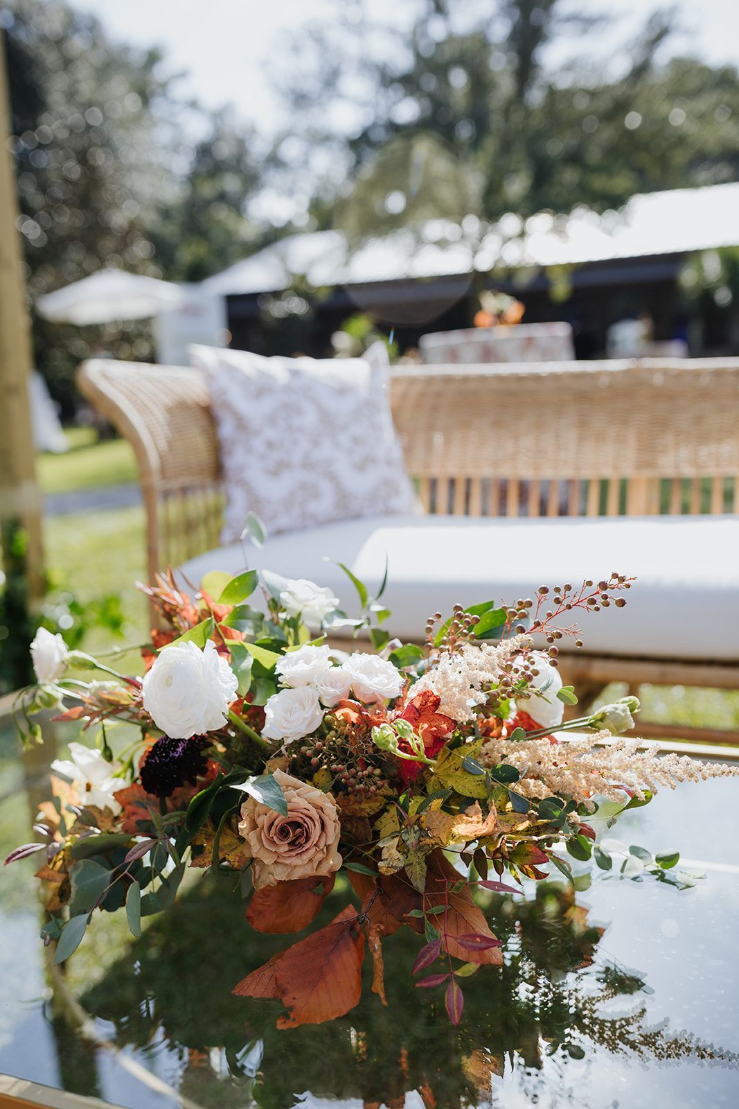 Floral arrangement on a glass table at an outdoor event with wicker chairs and white cushions in the background