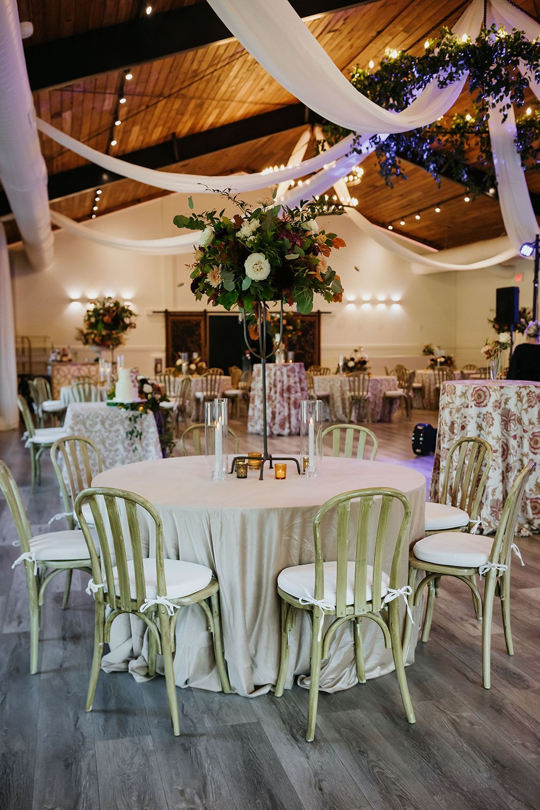Elegant event hall with round tables, white chairs, floral centerpieces, and draped string lights under a vaulted ceiling