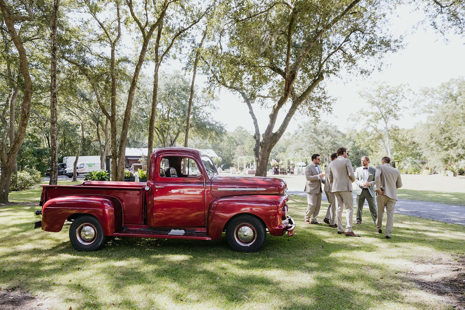 Red vintage pickup truck parked under trees with people standing beside it in a grassy field.